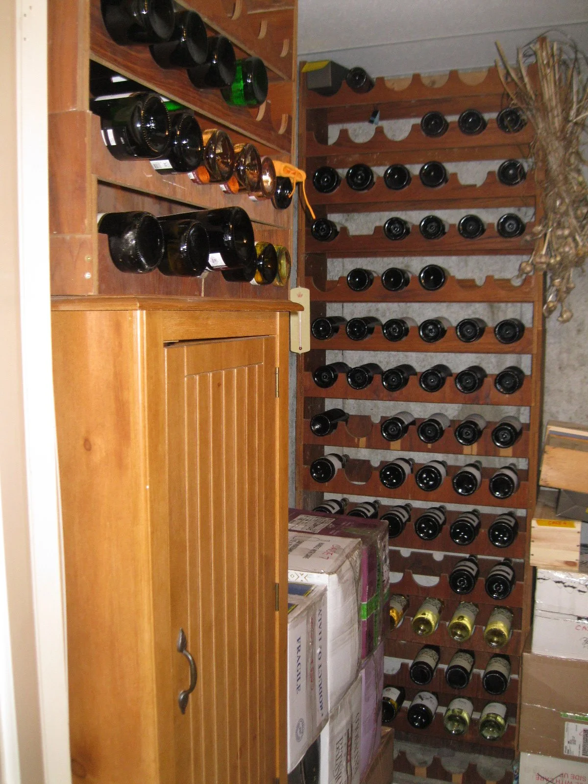 A wine cellar with wooden wine racks filled with bottles, a small wooden cabinet, and cardboard boxes stacked on the floor.