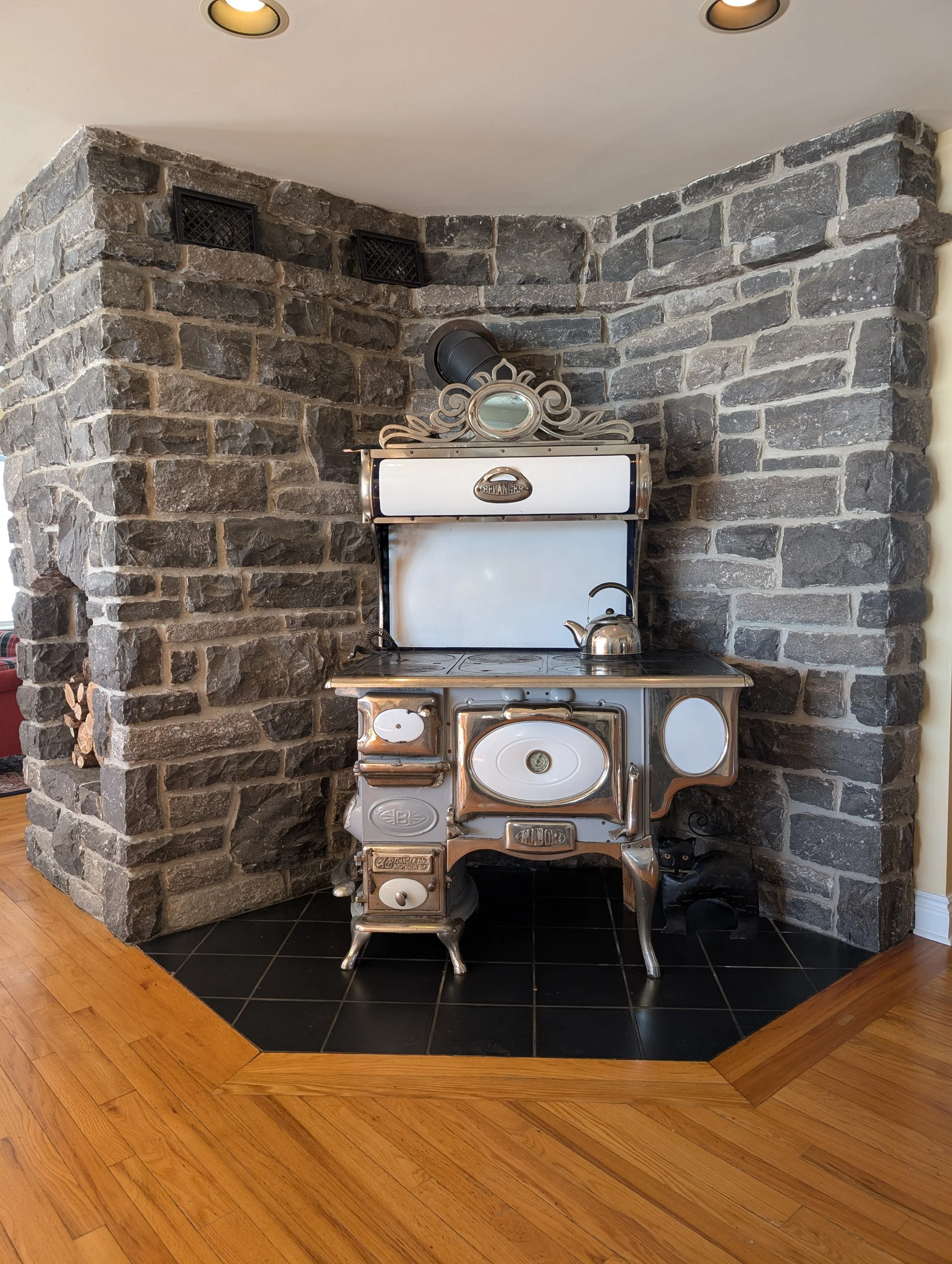 Vintage stainless steel stove with white accents against a gray stone wall, on a black tiled area with hardwood flooring nearby.
