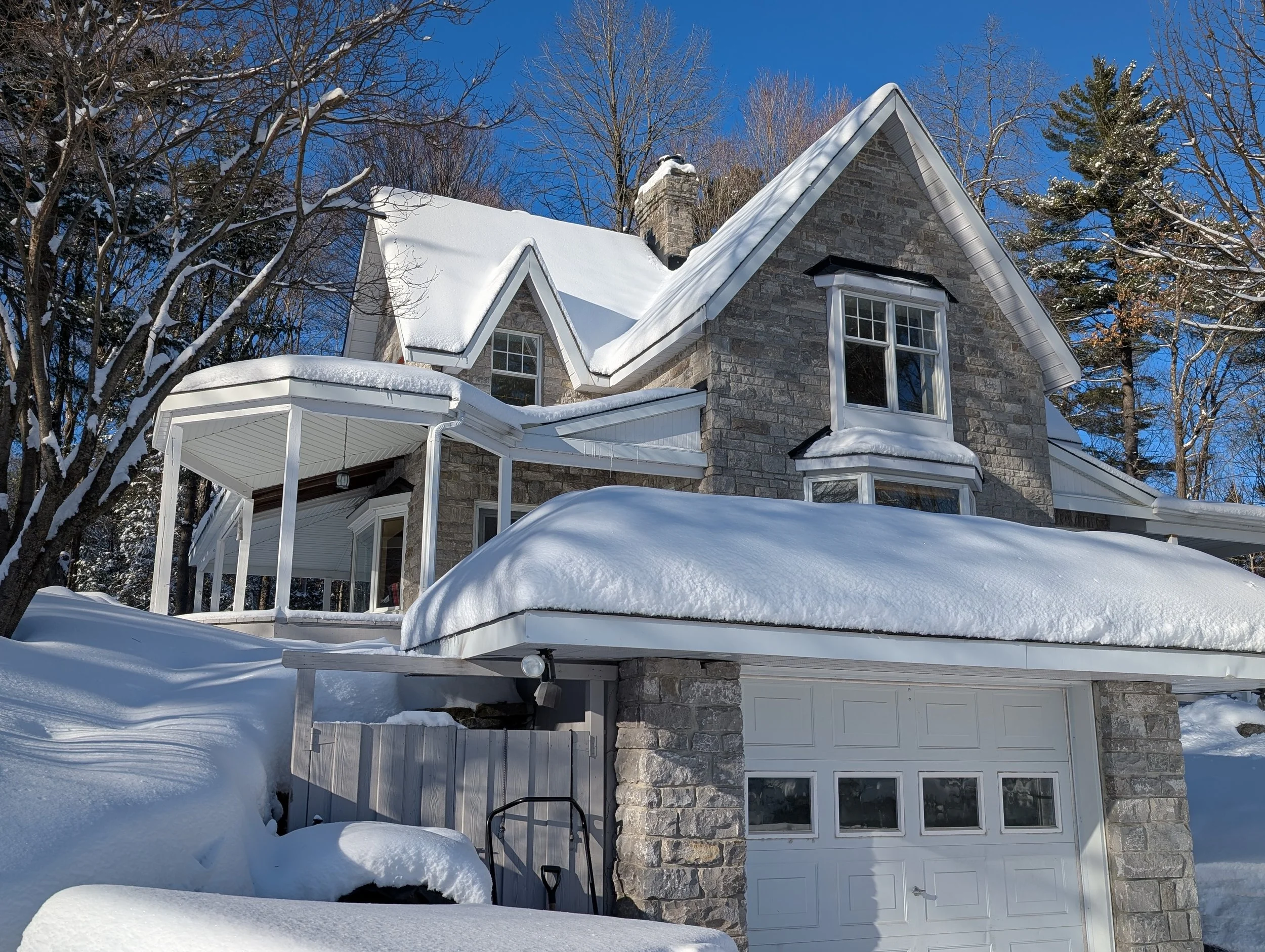 A large stone house covered in snow during winter, with a snow-covered roof, trees without leaves, and a blue sky in the background.