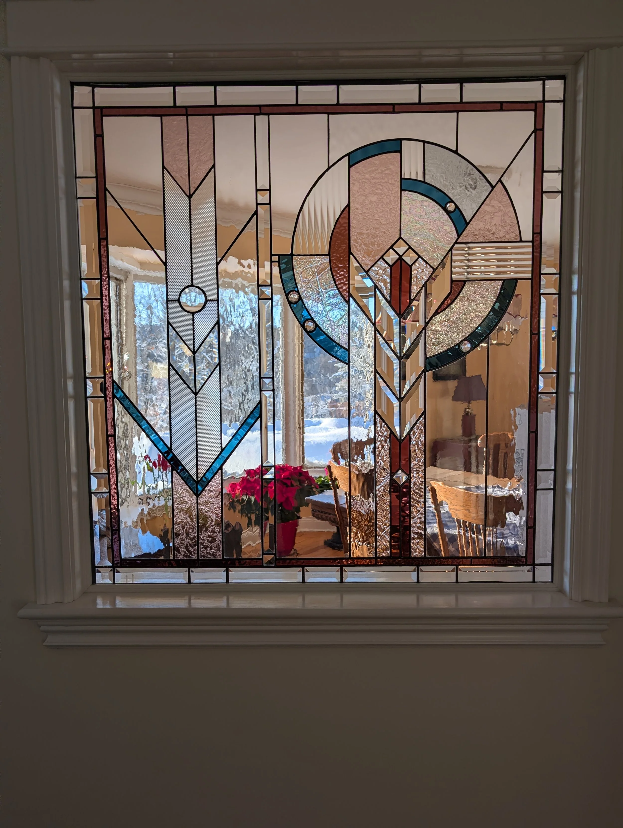 Decorative stained glass window showing geometric patterns with various colors, with a view of a snowy outdoor scene and a cozy indoor space with dining table, chairs, and potted poinsettia plant.