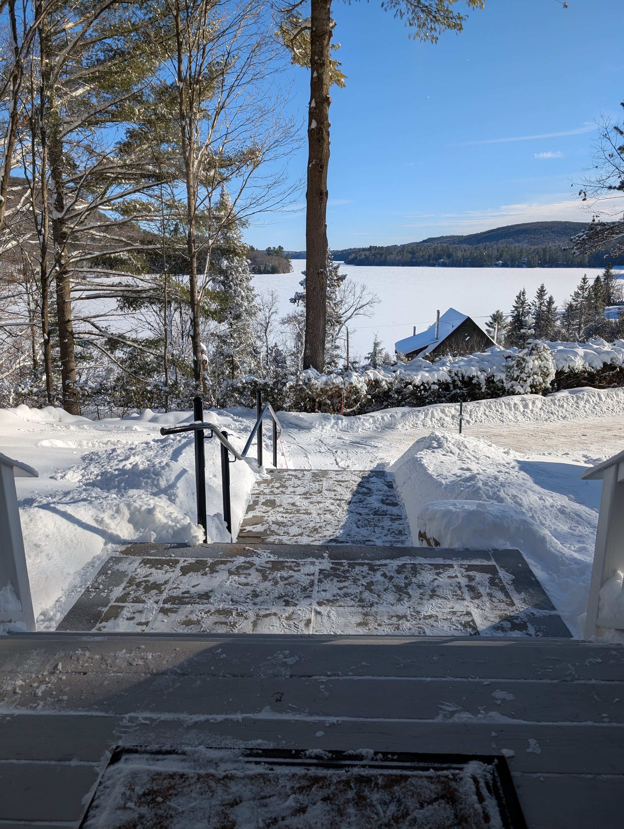 Snow-covered deck with steps leading down to a frozen lake, surrounded by snow-covered trees and a small house with a sloped roof, under a clear blue sky.