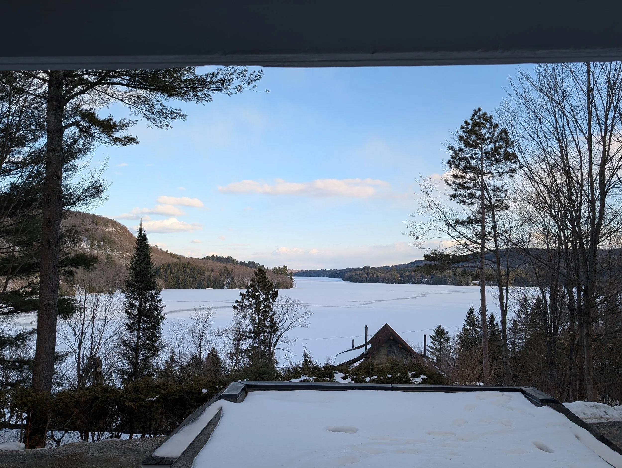 View of a frozen lake with snow, surrounded by trees and hills under a partly cloudy sky, seen from a porch or balcony with a snow-covered roof in the foreground.