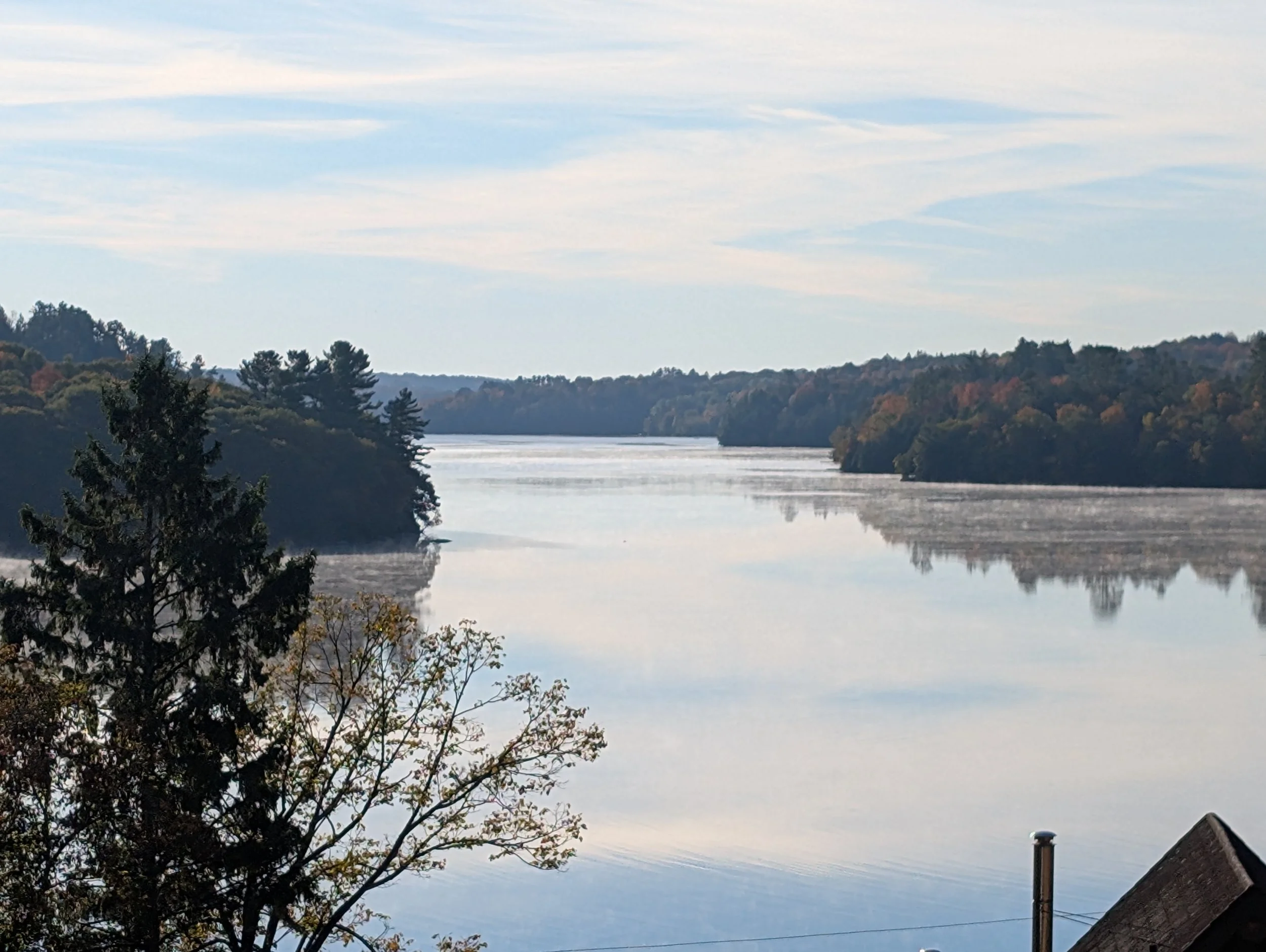 A calm river reflecting the sky and trees along its banks, with some clouds and autumn foliage.