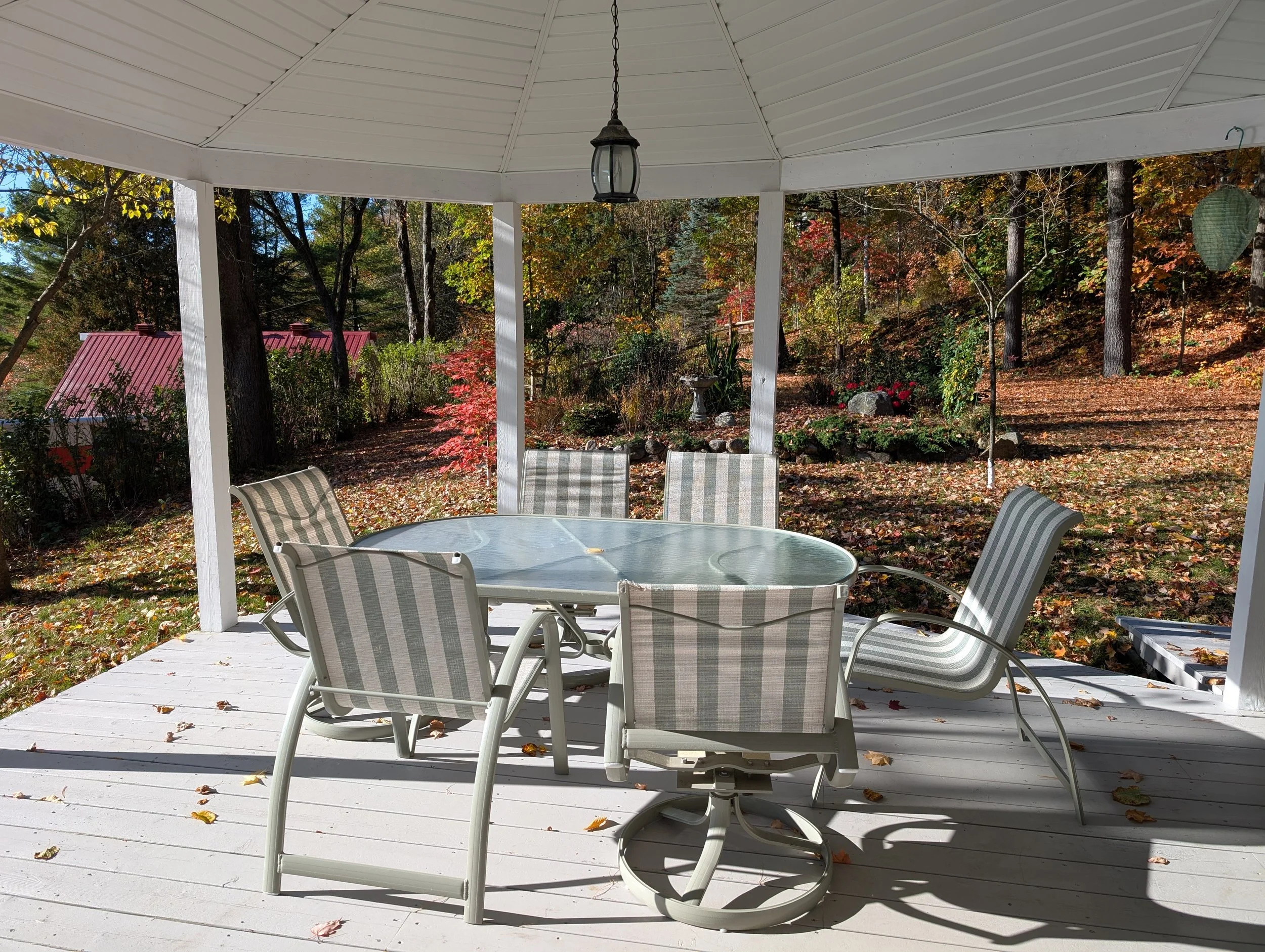 Outdoor patio with a round glass table surrounded by six striped patio chairs on a wooden deck, with a background of trees and fall foliage.
