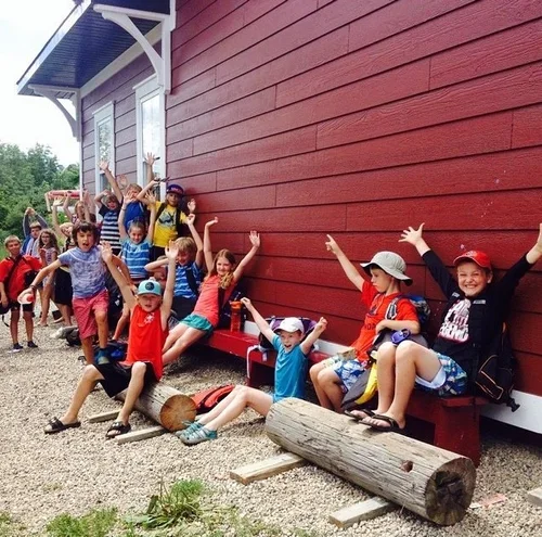 Group of children and adults posing happily outdoors next to a large red wooden building.