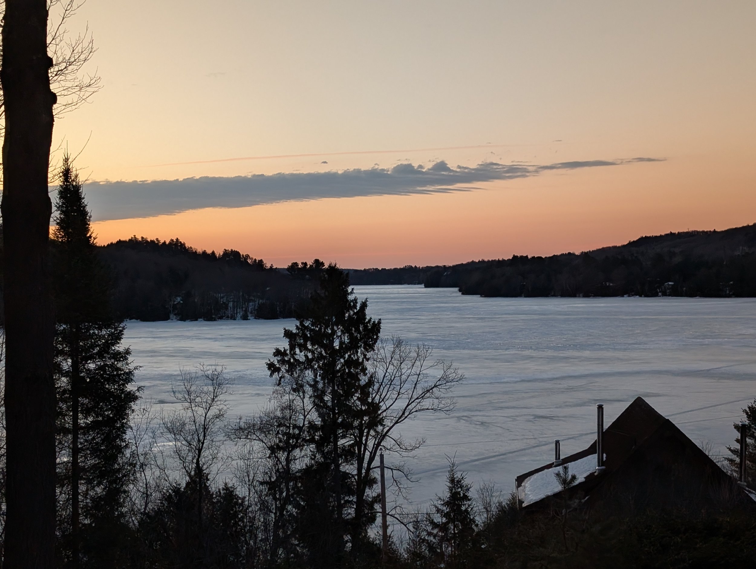 Snow-covered frozen lake at sunset with trees and a house silhouette in foreground, hills in background.