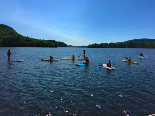 People paddleboarding on a lake with wooded hills in the background under a blue sky.