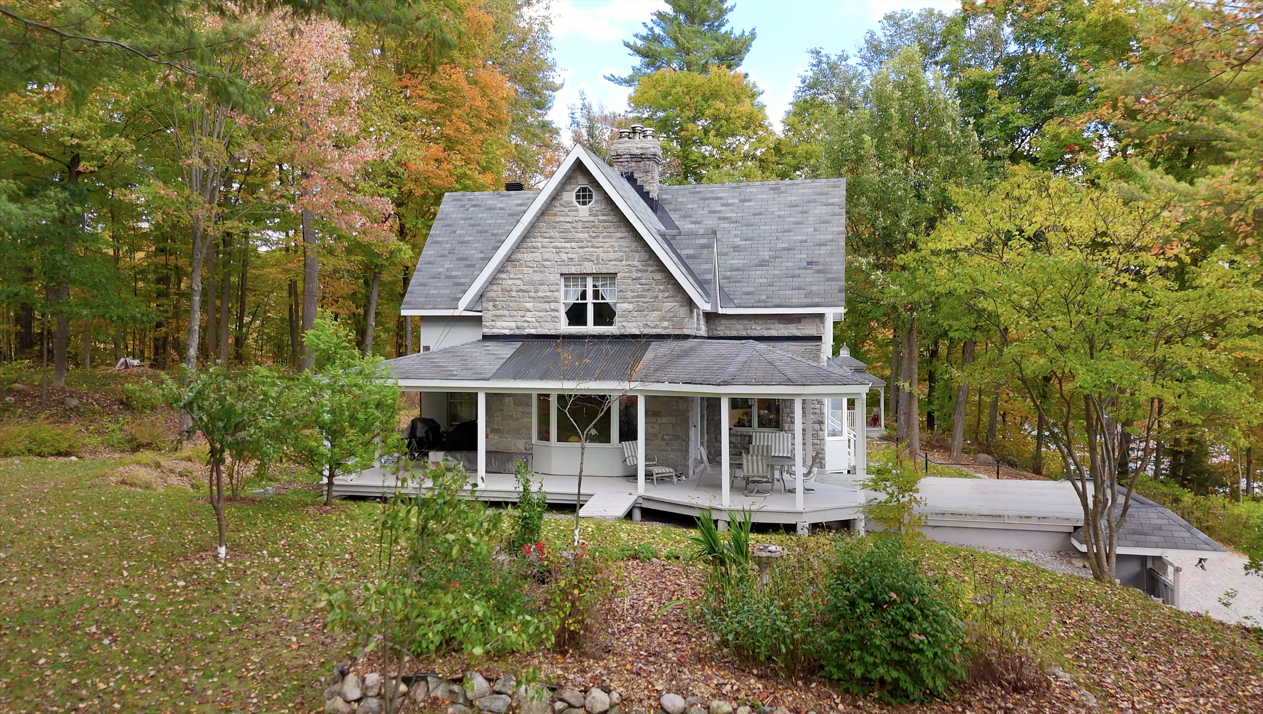 A two-story house with a stone facade and gray shingle roof, surrounded by trees with autumn foliage, and a wrap-around porch with outdoor furniture.
