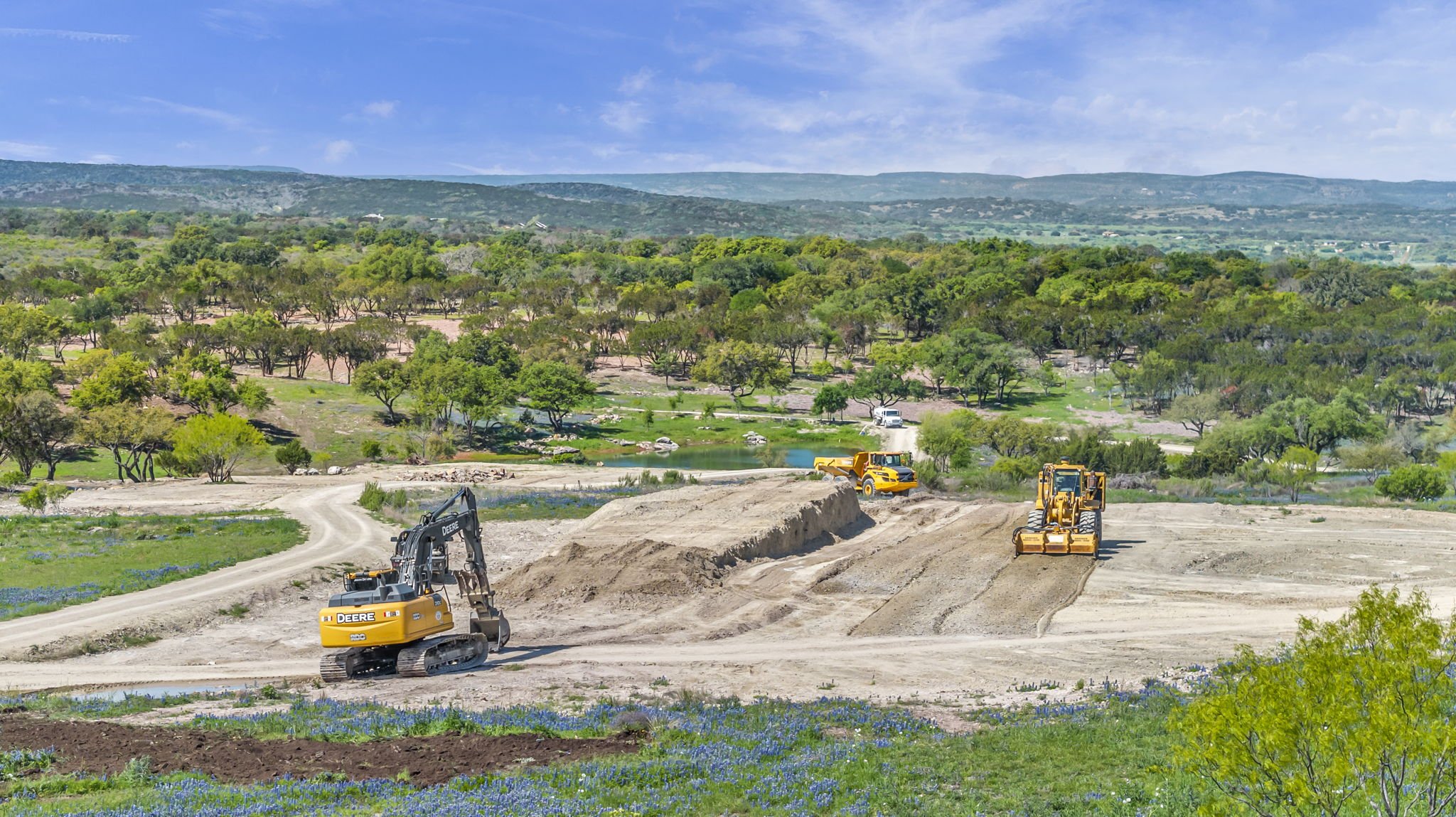 Construction site with yellow bulldozers and excavators on dirt, surrounded by greenery, trees, and hills under a partly cloudy sky.