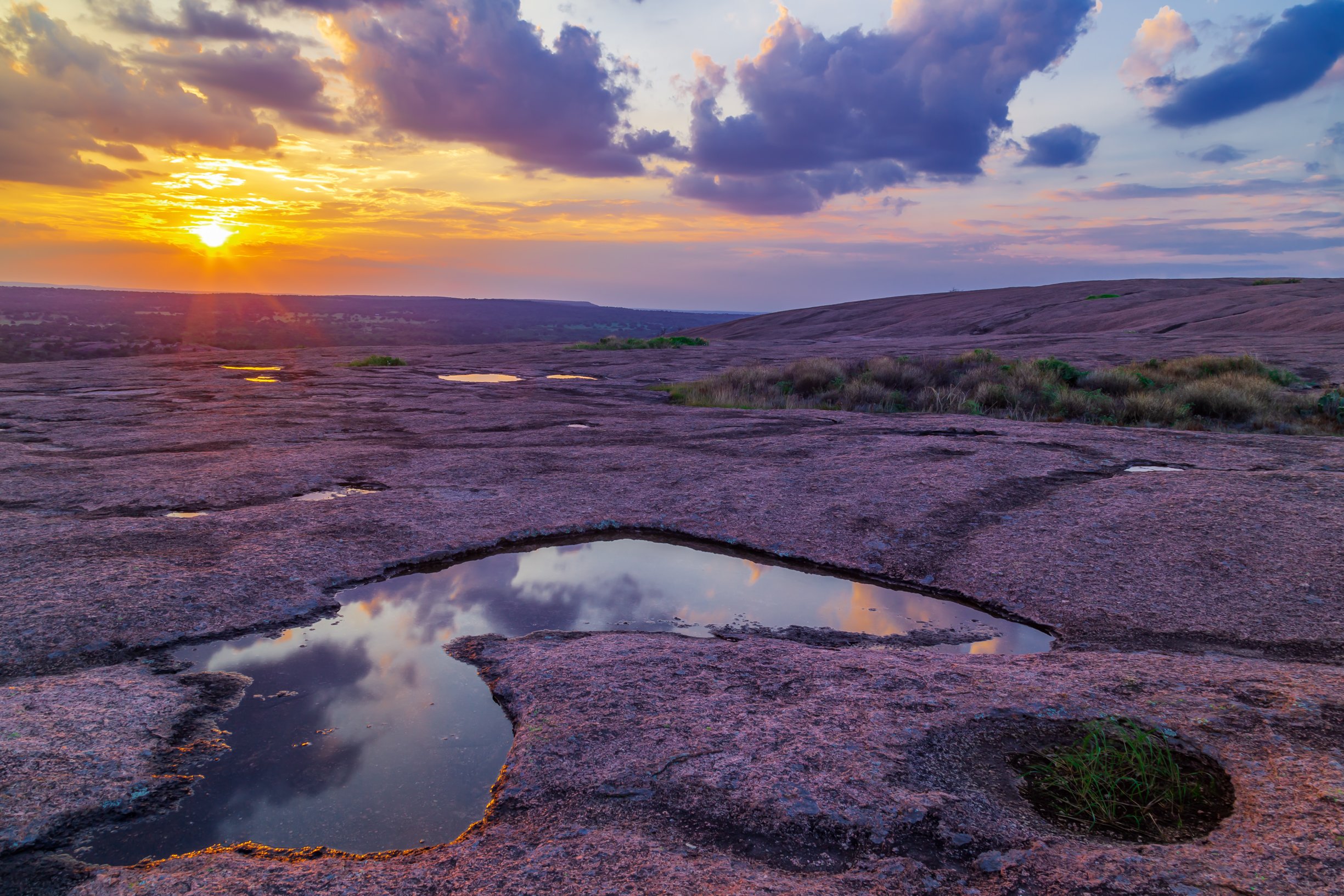 Sunset over a rocky landscape with small pools of water reflecting the sky and clouds.