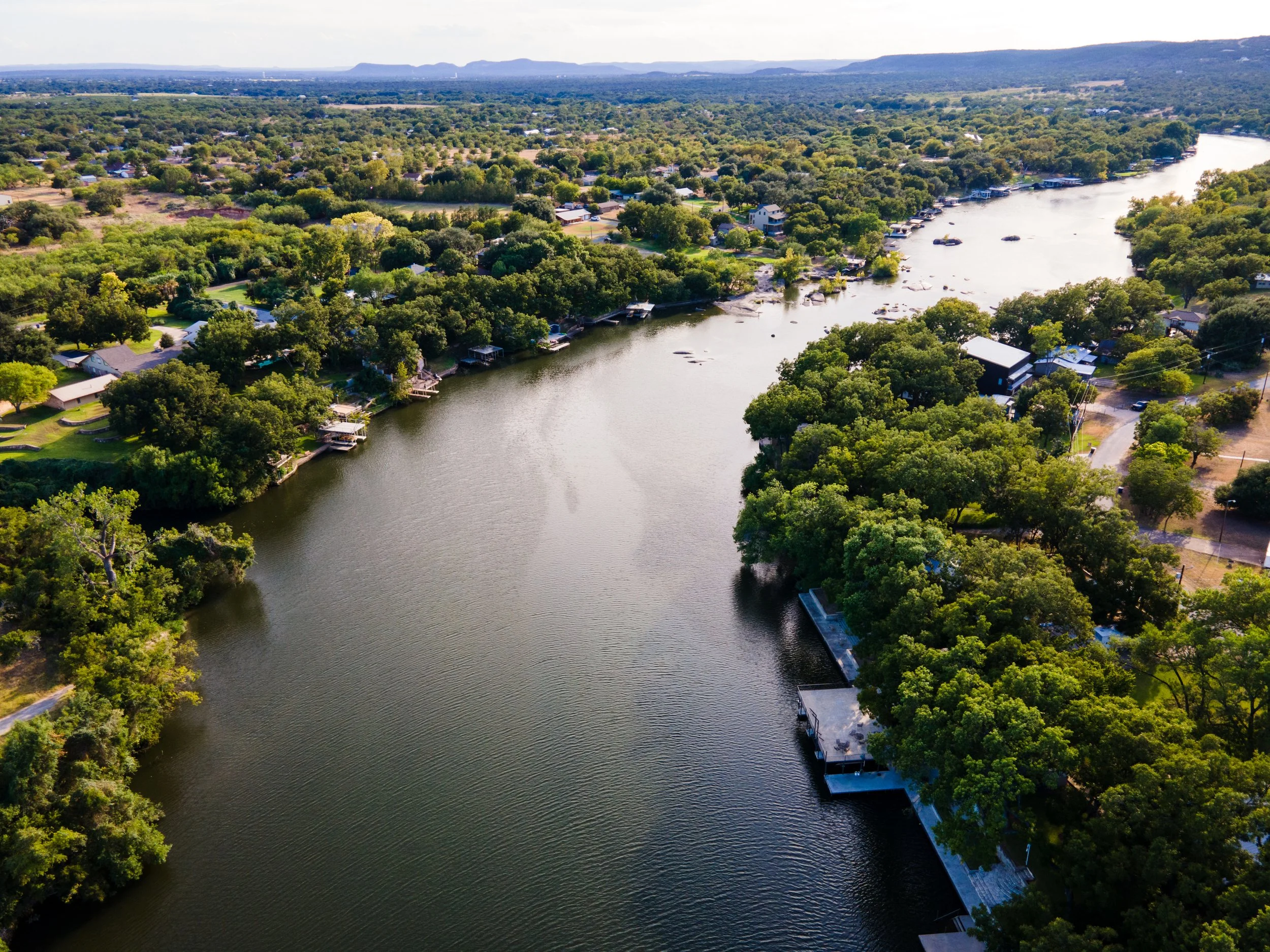 Aerial view of a river winding through a lush, green town with houses along the shoreline and boats docked nearby, surrounded by trees and a distant mountain range.