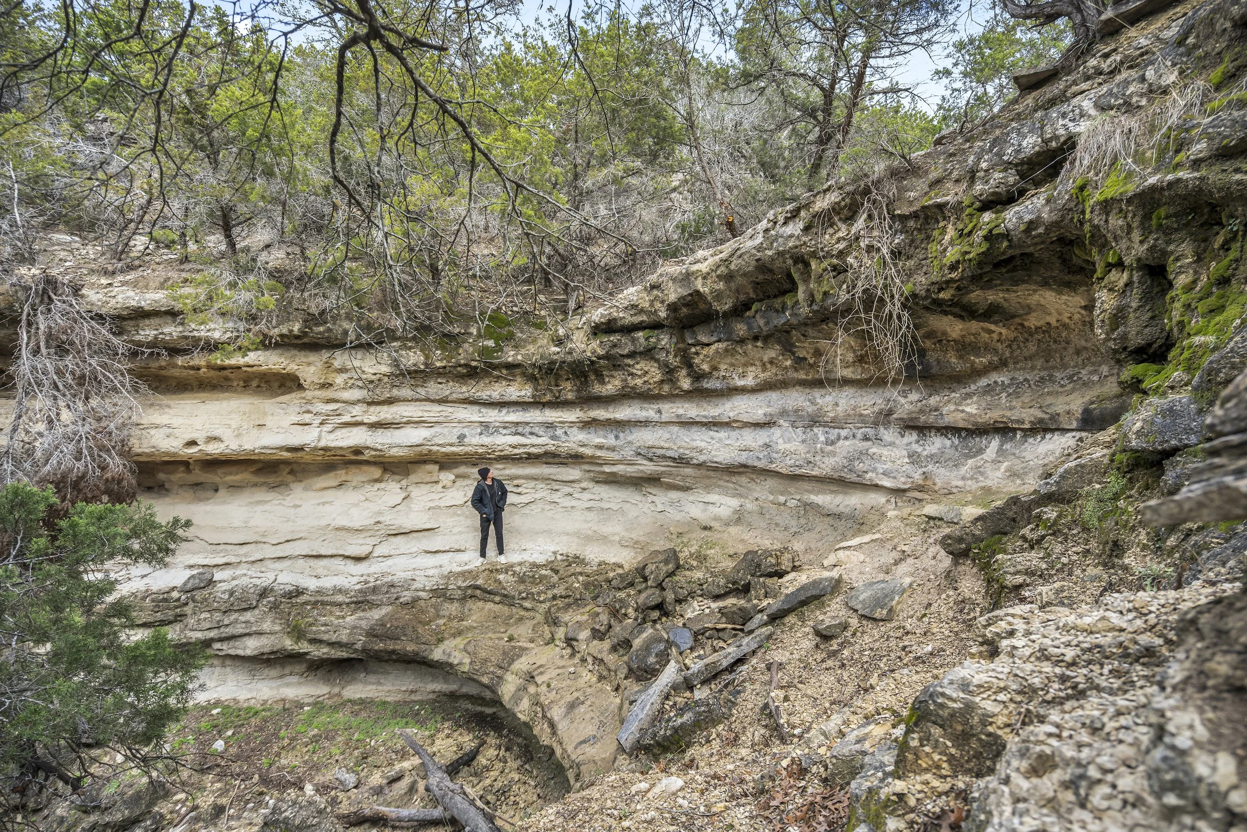 Person standing near layered rock formations and overhanging cliffs in a wooded area with green trees.