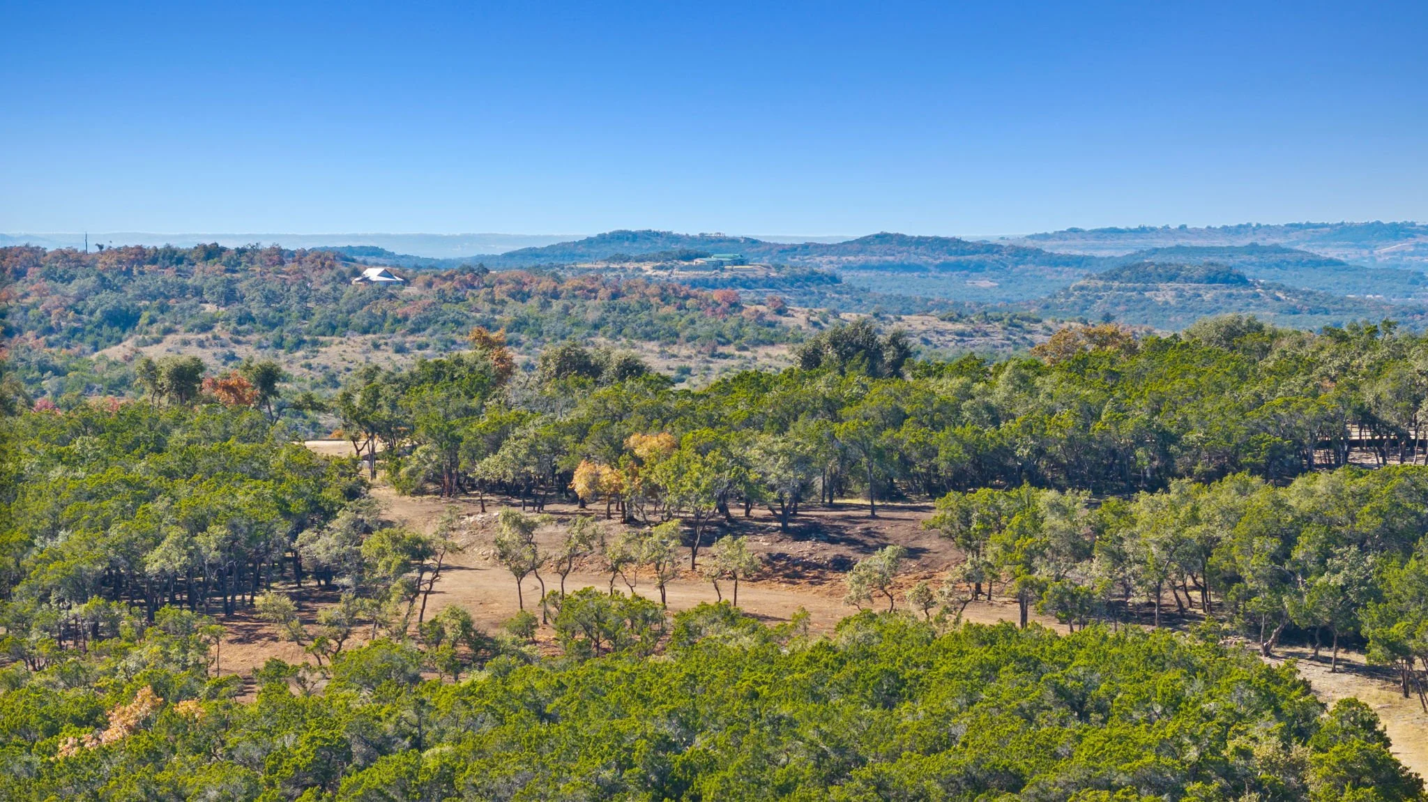 A scenic view of rolling hills at RanchesAt Dripping Springs covered in green and brown trees under a clear blue sky.