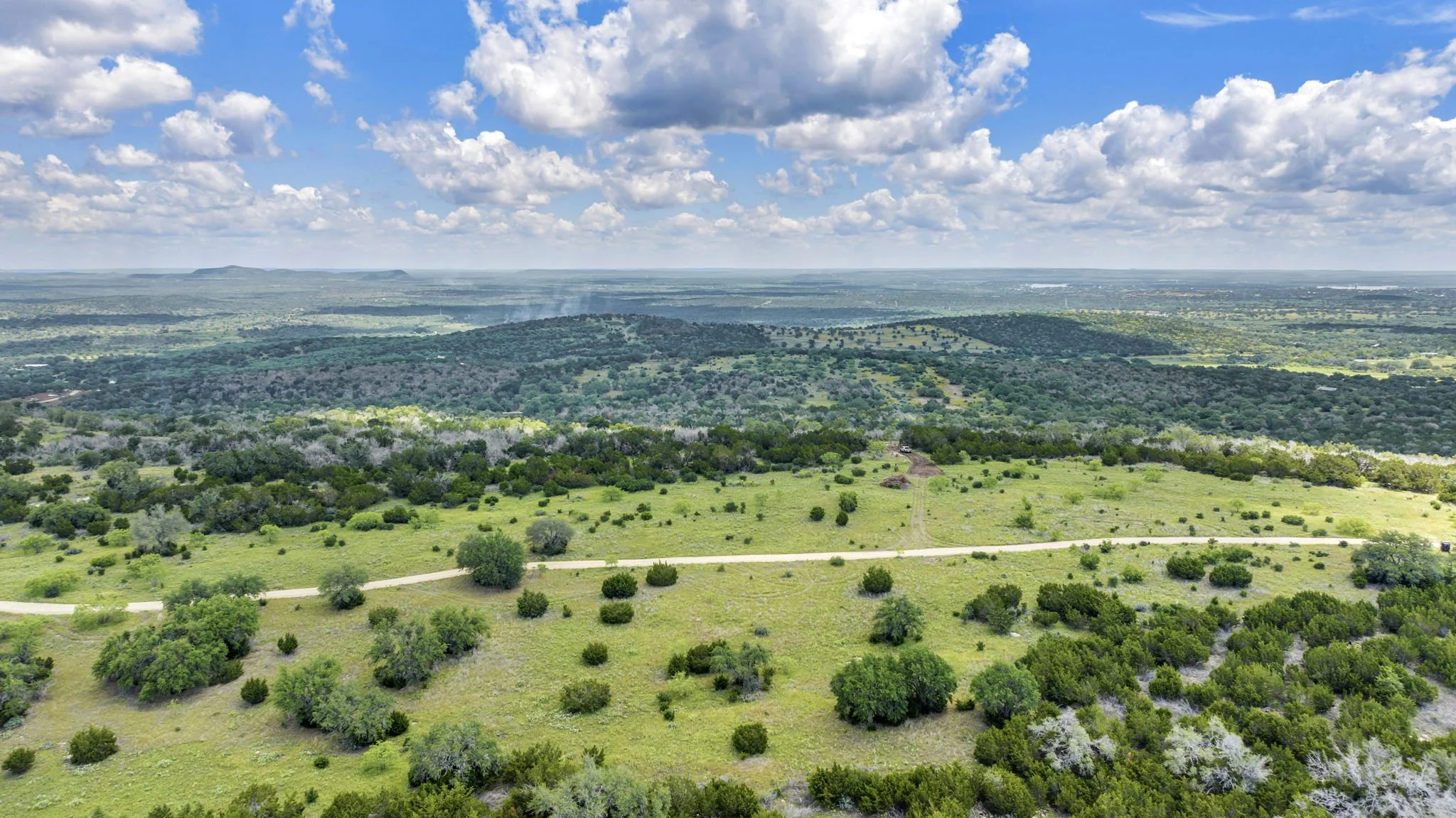 Aerial view of a lush green landscape with rolling hills, scattered trees, panoramic views, and a dirt road winding through the terrain under a partly cloudy sky.