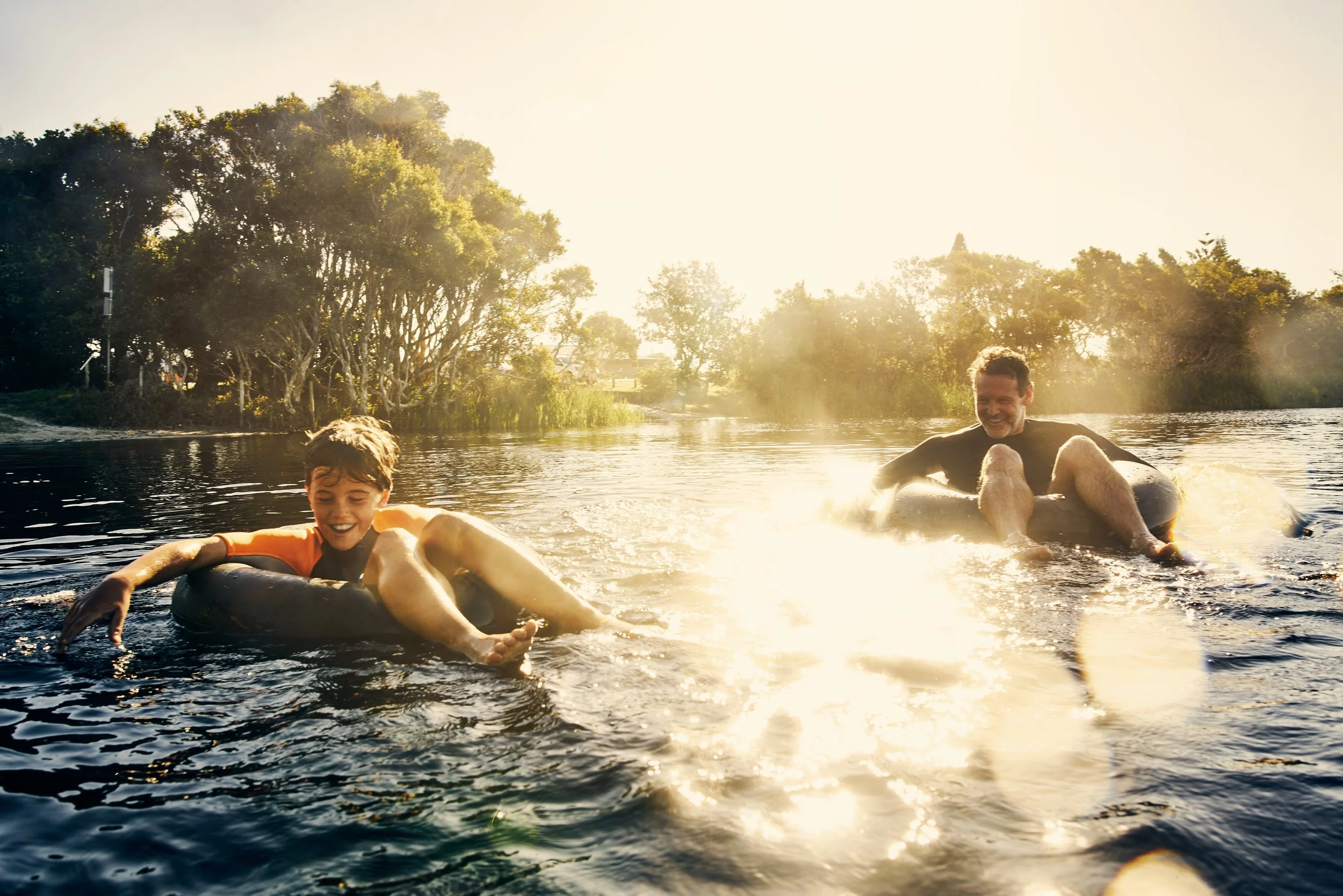 Two people, a young boy and an adult, riding inner tubes on a river at sunset, surrounded by trees.