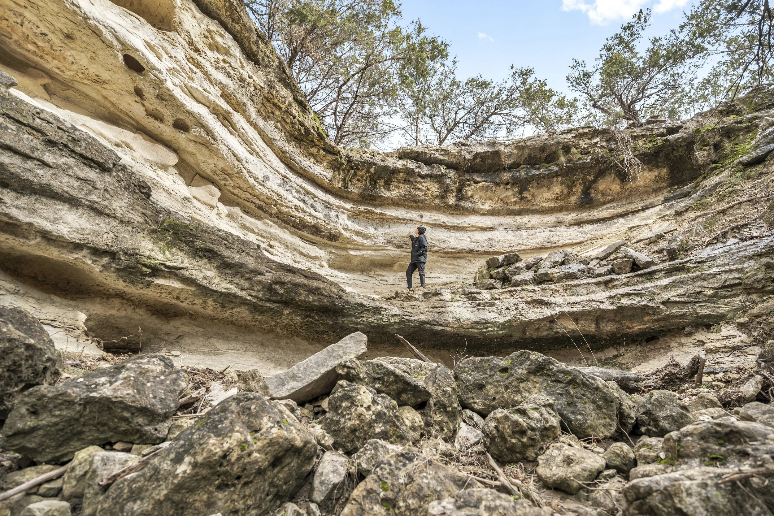 A person standing on a rocky ledge in a large rock formation with layered sedimentary rock, trees in the background, and a clear sky above.