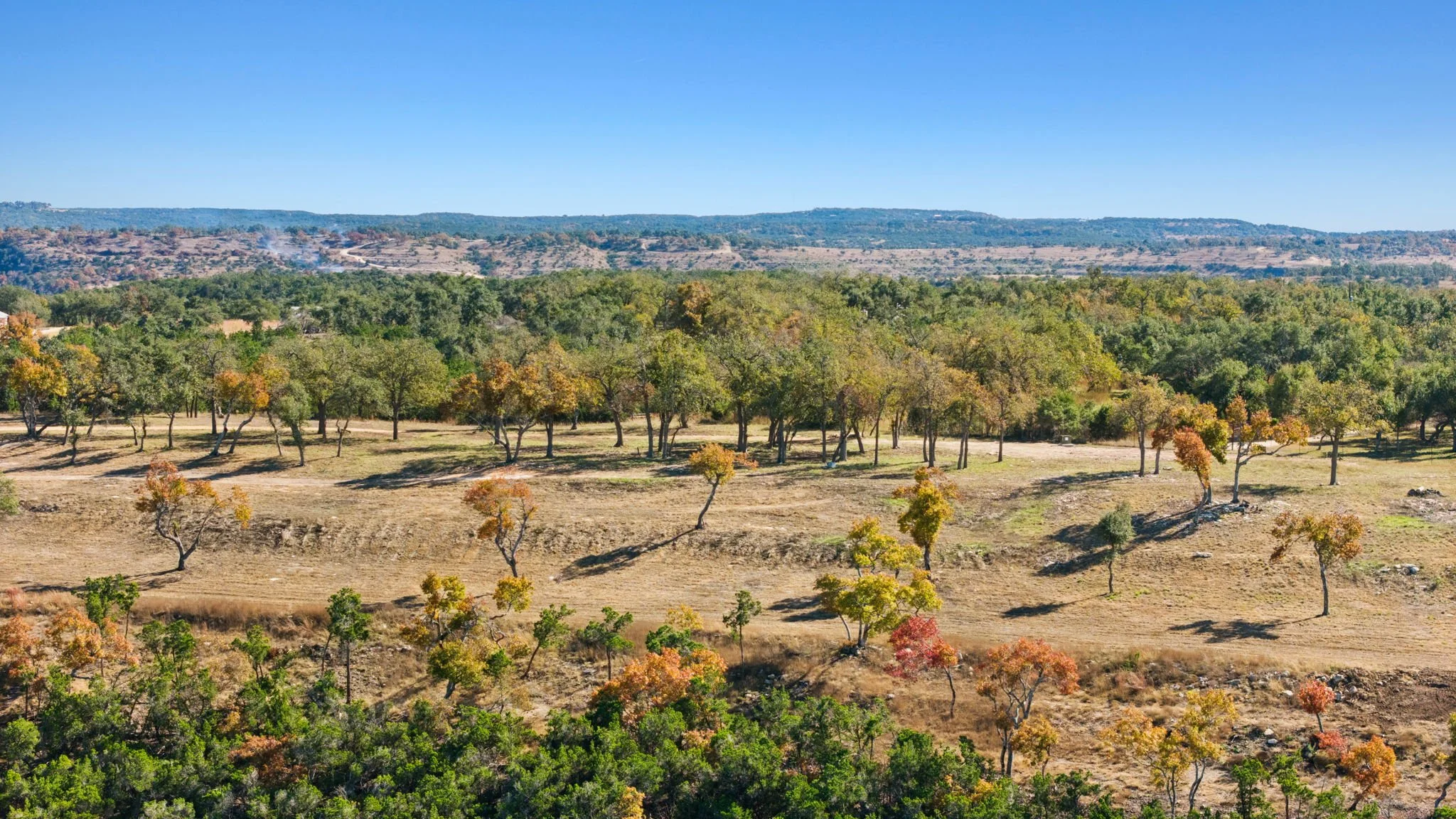 Scenic landscape of RanchesAt Dripping Springs displaying an open field with scattered trees displaying autumn foliage, hills in the distance, and a clear blue sky.