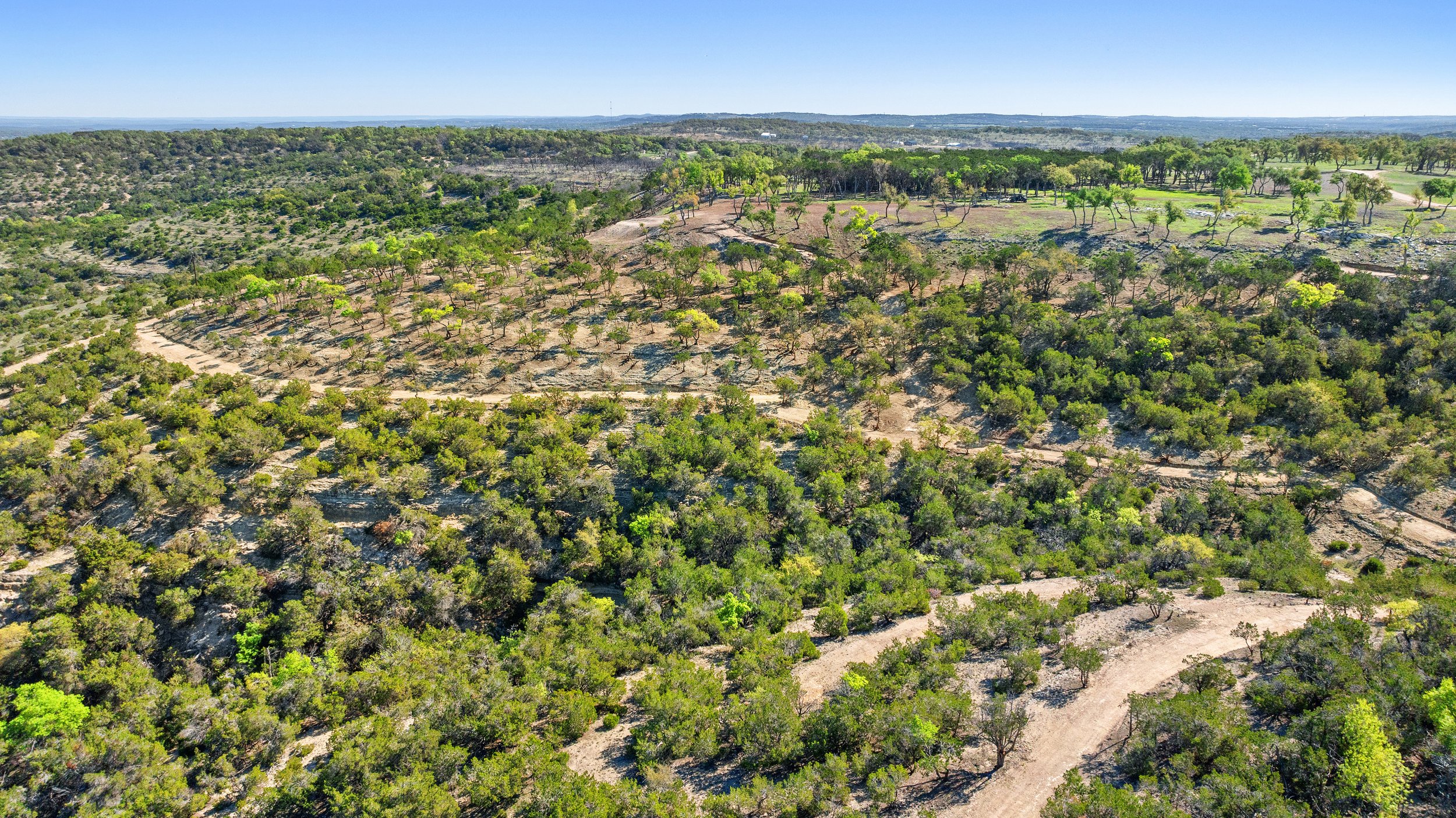 Aerial view of a rural landscape with dirt roads, green trees, and hilly terrain under a clear blue sky.