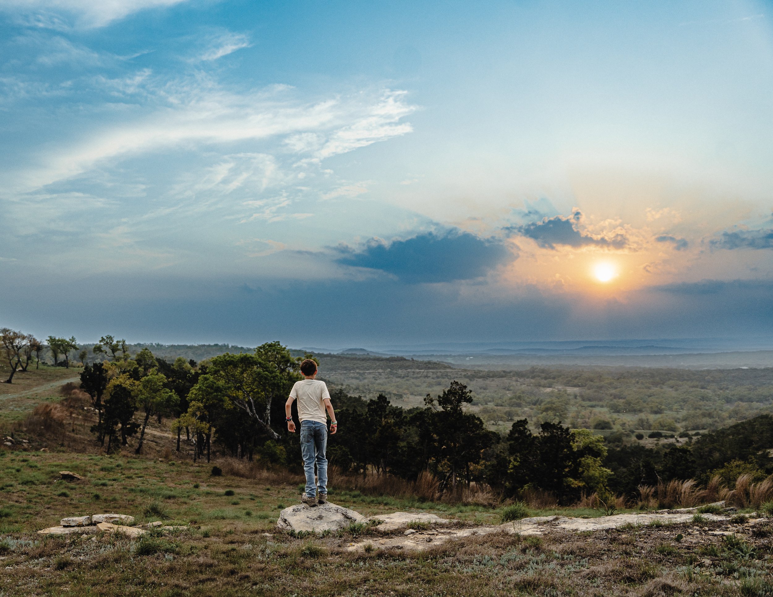 A person standing on a rock in a grassy field, facing the iconic Texas Hill Country landscape with trees, hills, and a sunset in the distance.