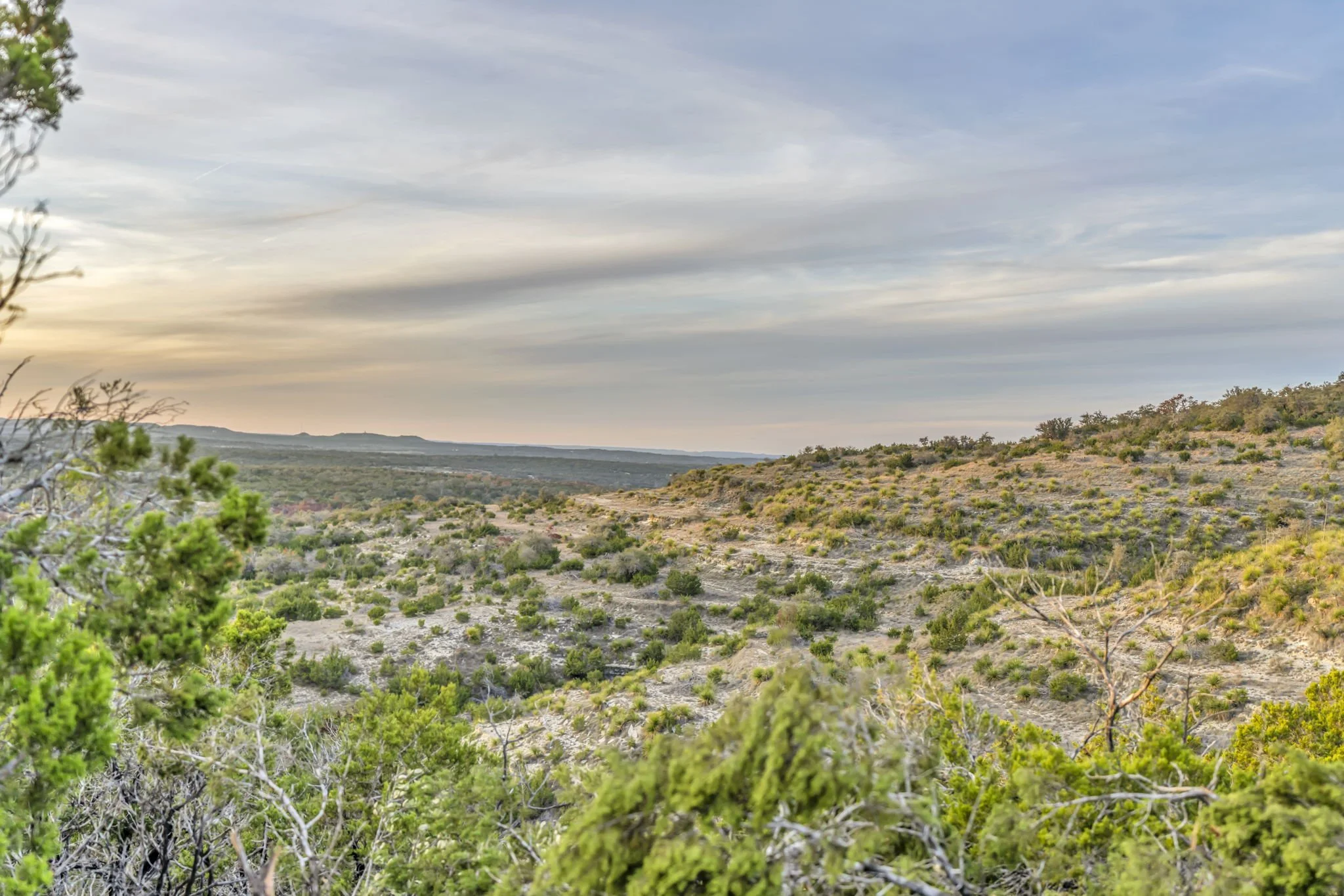 A scenic landscape with rolling hills covered in sparse green shrubs and trees. The sky is cloudy with soft sunset lighting.