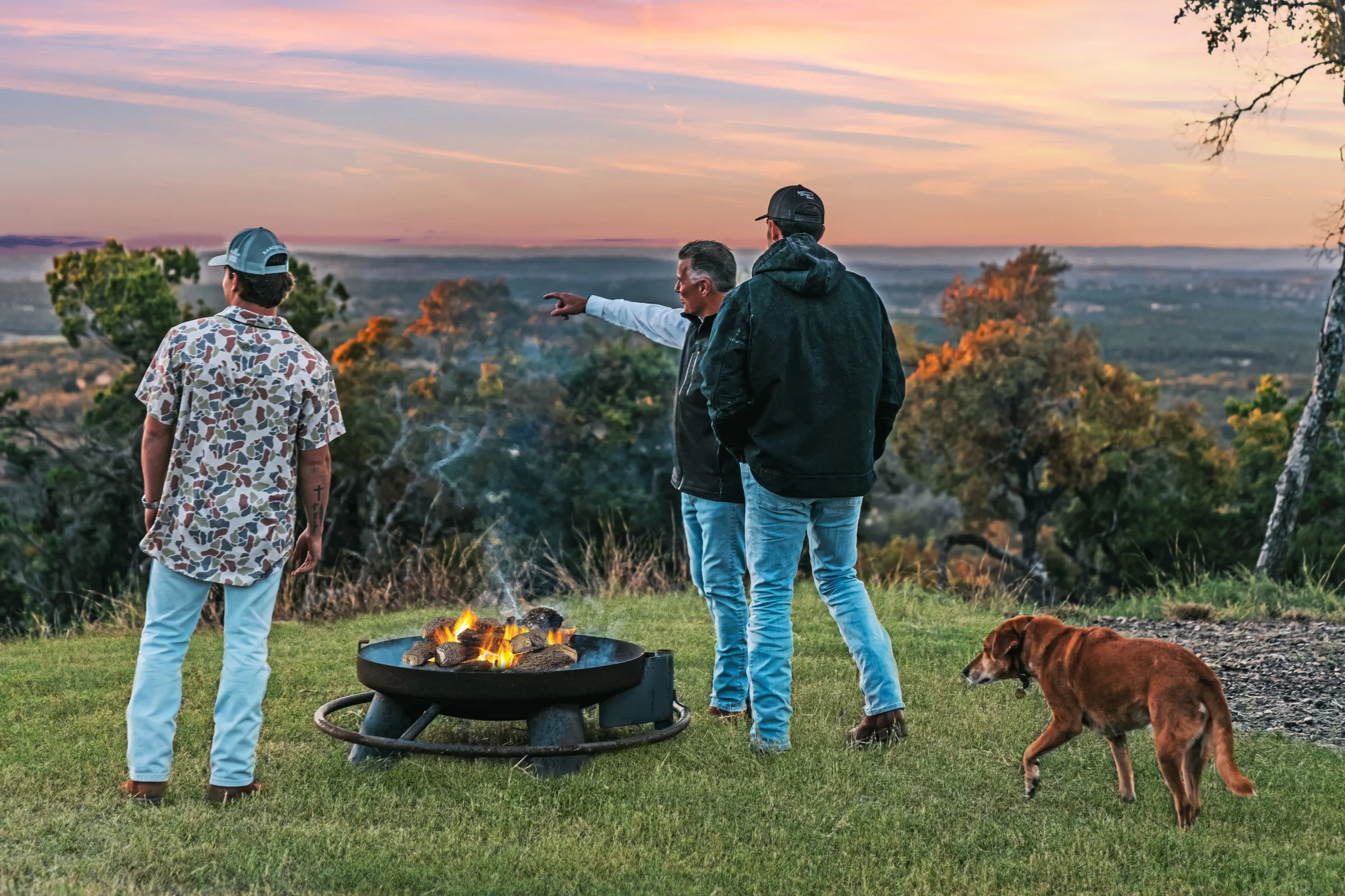 Three men and a dog standing on a grassy hill near a fire pit with a scenic view of trees and the sunset in the background.