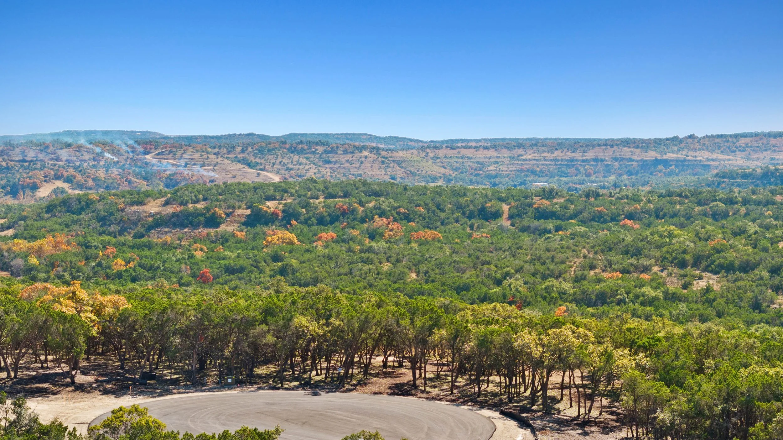 A wide view of a landscape with a circular dirt area at the front, a dense forest of green trees, and rolling hills with some smoke and a winding road under a clear blue sky.