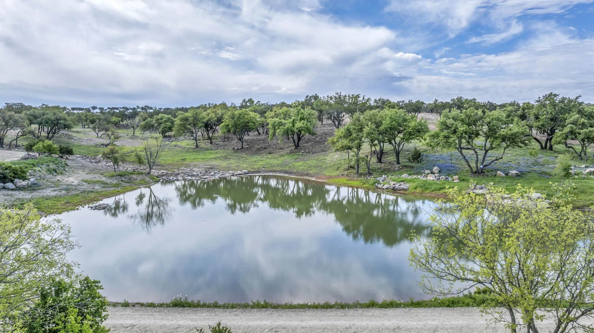A landscape with a pond reflecting the sky and trees, surrounded by greenery, rocks, and hills under a partly cloudy sky.