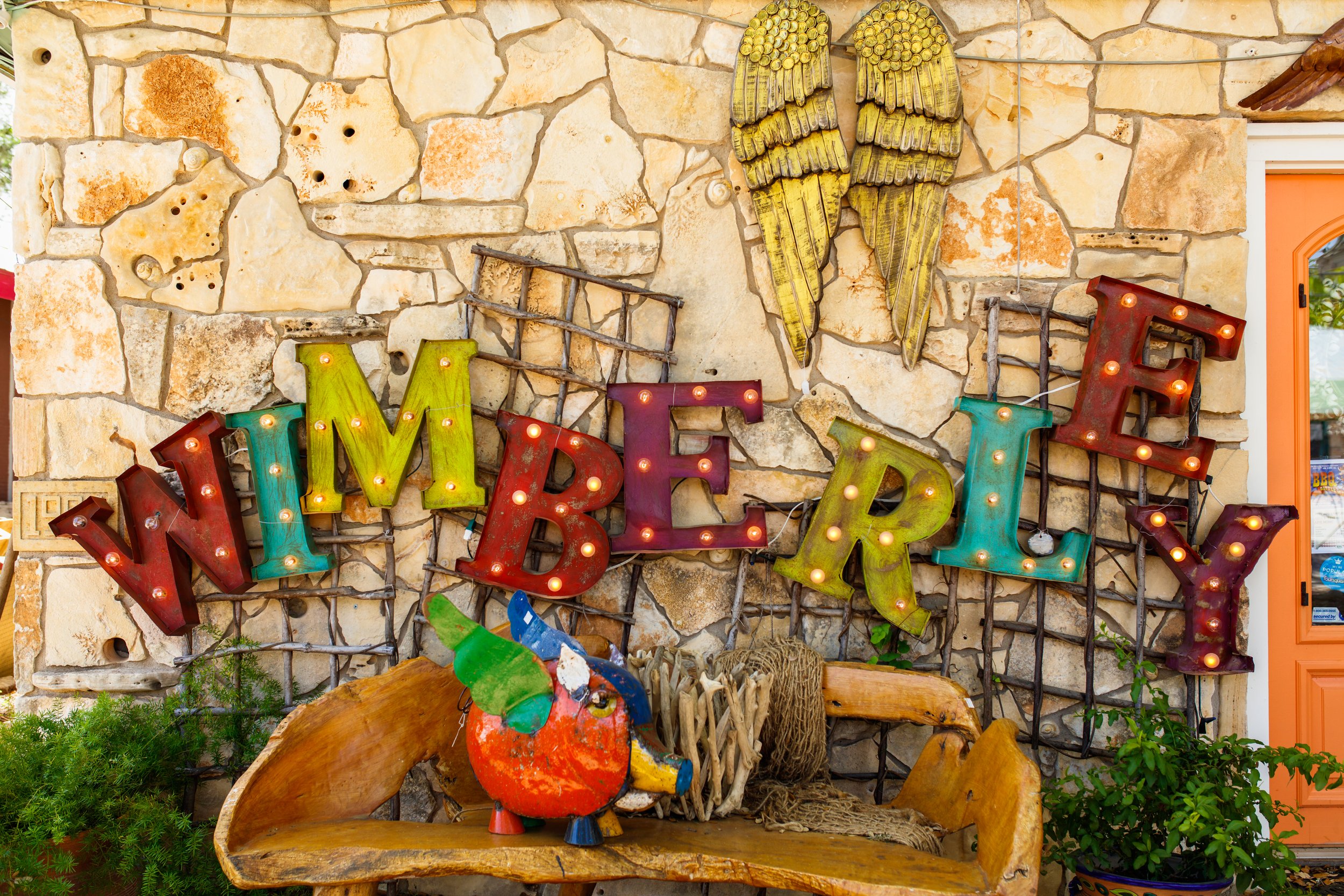 Decorative sign that reads 'WELCOME' with colorful, illuminated letters, mounted on a stone wall alongside a metal angel wing sculpture, a rustic parrot figurine, and greenery, with a wooden bench in front.