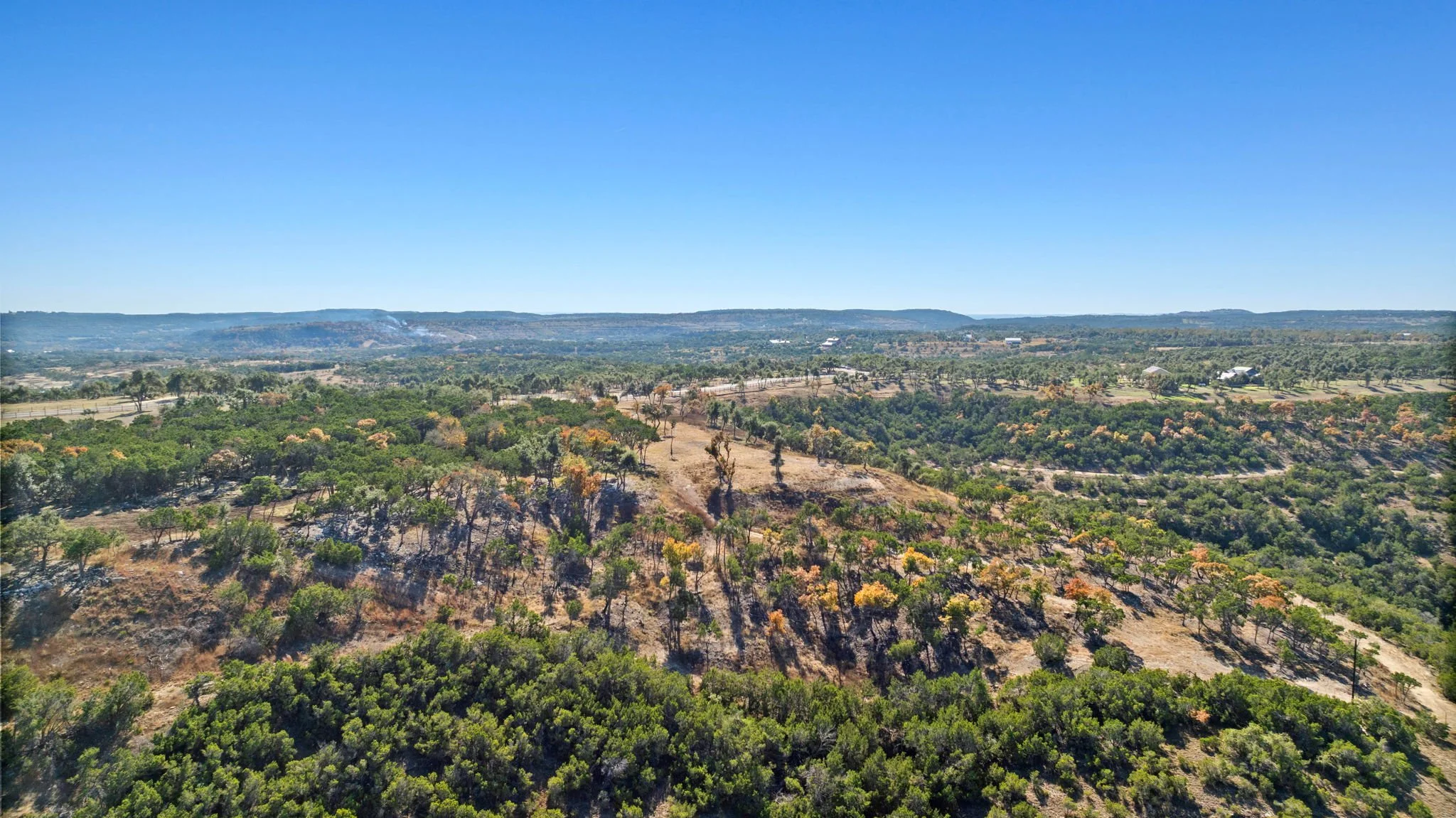 Aerial view of a hilly landscape with scattered trees, some trees appear to be burning, smoke rising in the distance, clear blue sky overhead.