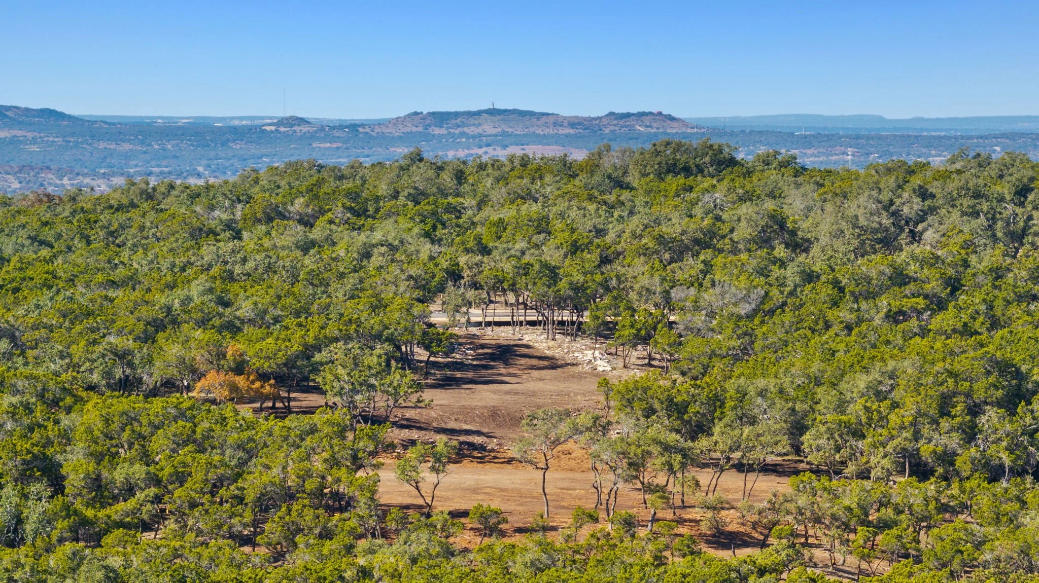 A scenic view of a lush green forest with patches of cleared land, and hills in the distance under a clear blue sky.