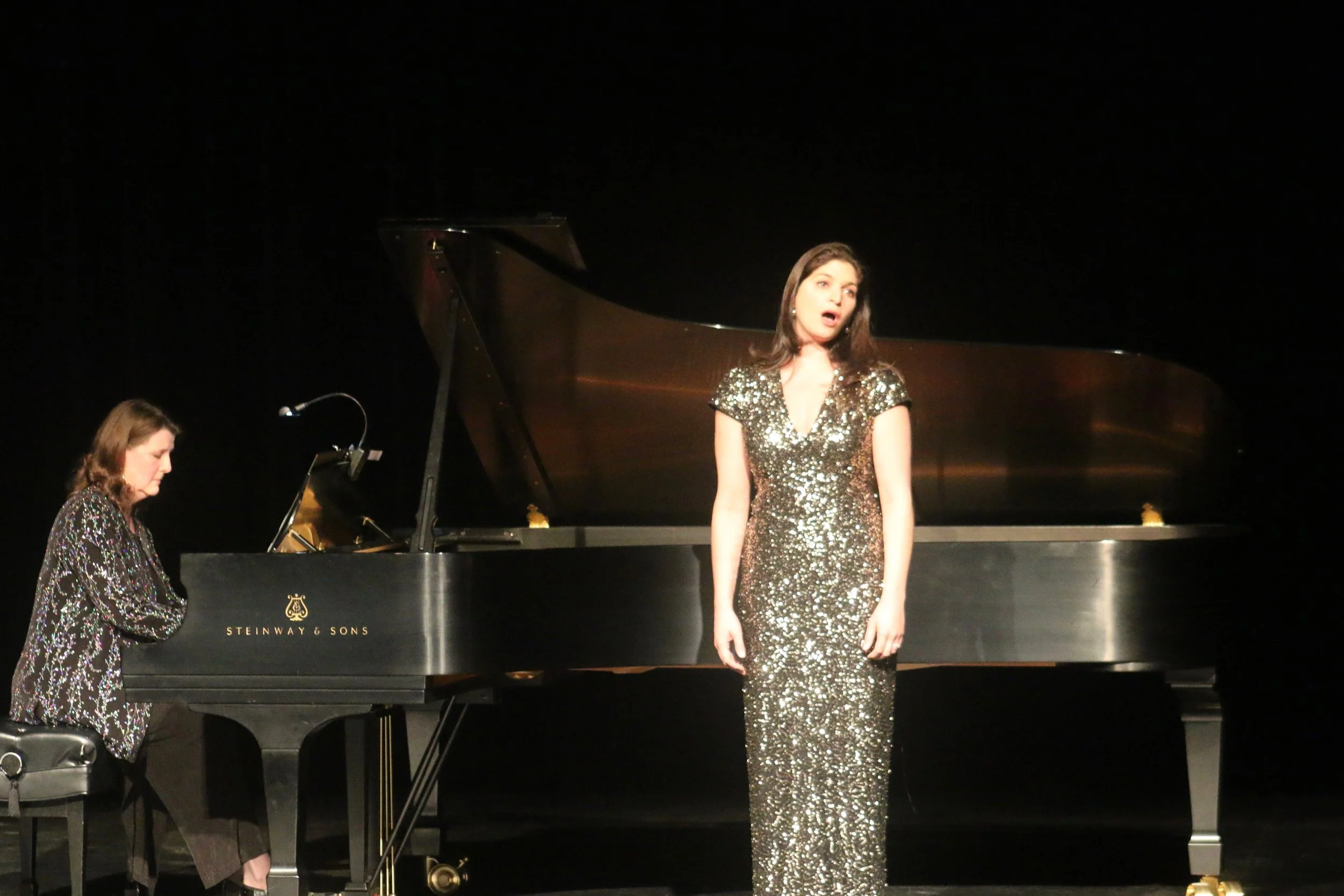 Dr. Victoria Moss, board certified dermatologist in a sparkly gold gown stands singing an operatic aria in front of a black piano on a stage, while a pianist plays nearby.