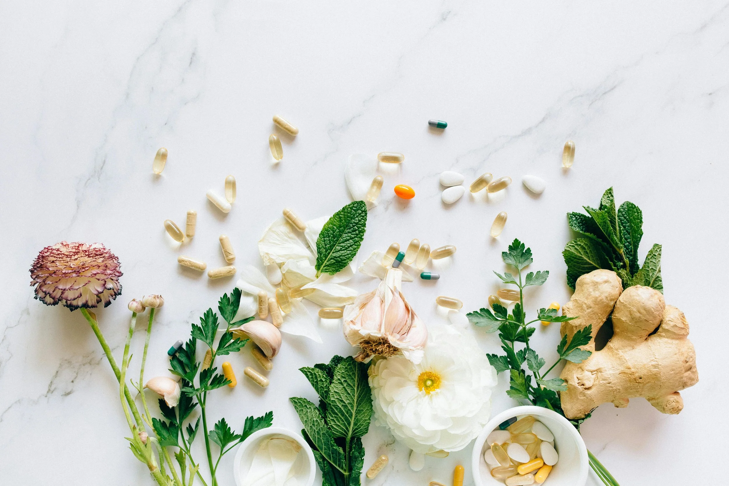 A flat lay of herbs, flowers, garlic, ginger, and various pills and supplements on a white marble surface.
