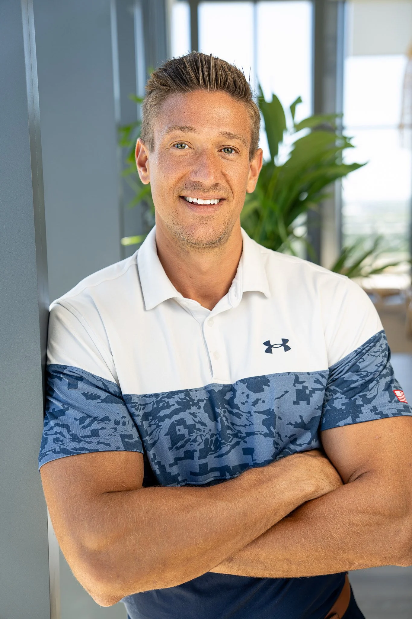 A smiling man standing indoors with arms crossed, wearing a white and blue Under Armour polo shirt, with a plant and windows in the background.