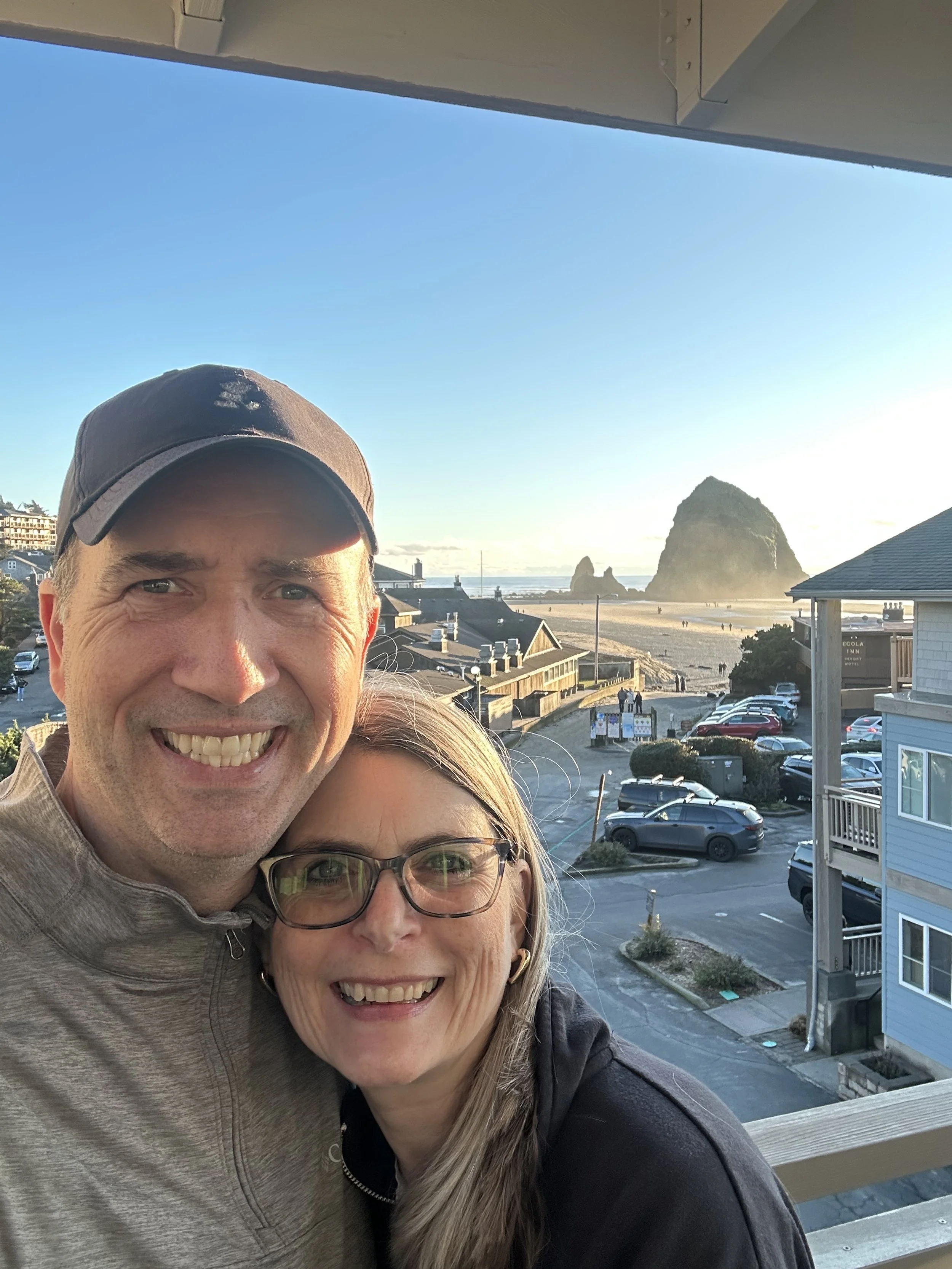 A smiling man and woman taking a selfie on a balcony overlooking a beach with rock formations, houses, and parking lot in the background.