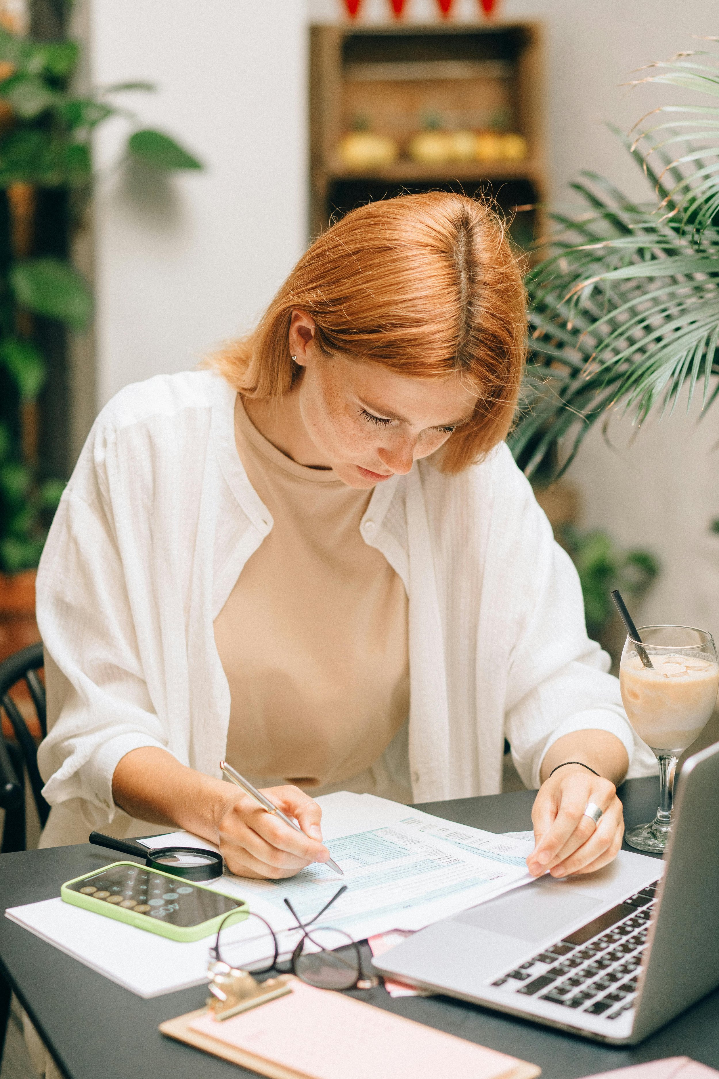 A young woman with red hair and freckles working at a desk, writing on papers with a pen. The desk has a smartphone, glasses, and a laptop. There is a glass of iced coffee or latte beside her, and green plants in the background.