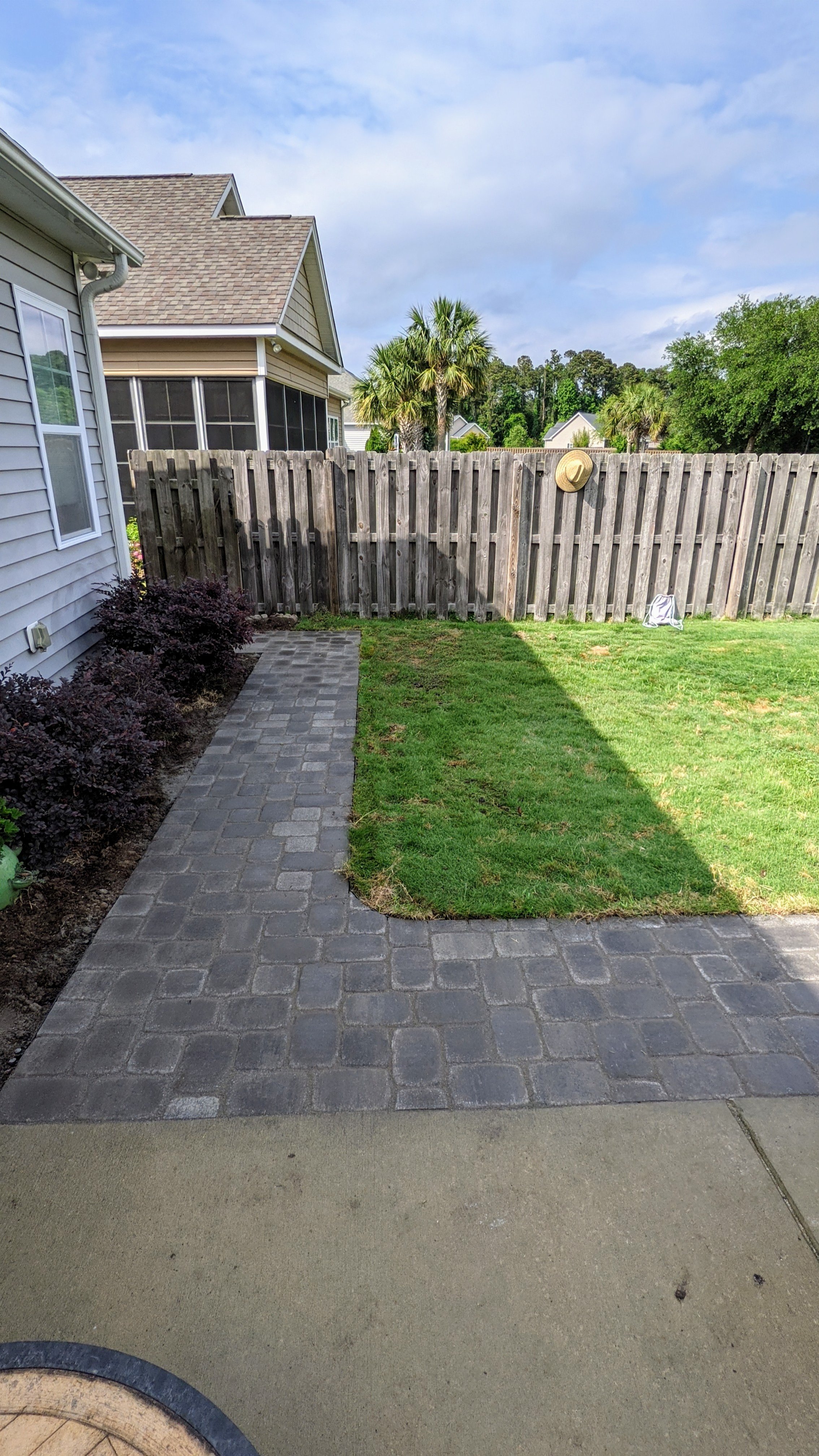 A backyard with a brick walkway, a patch of grass, a wooden fence, and some bushes. There are neighboring houses and trees in the background under a partly cloudy sky.