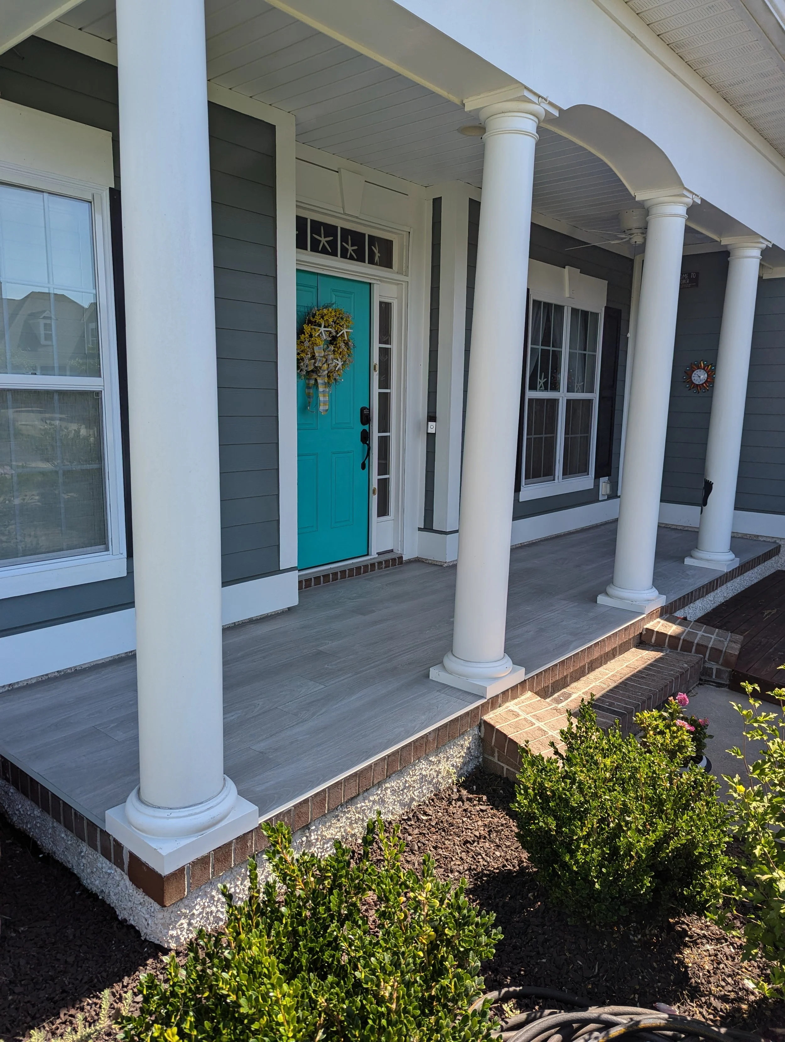 Front porch of a house with a teal door, white columns, and a wreath on the door. There are bushes and plants in front, and brick steps leading to the porch.