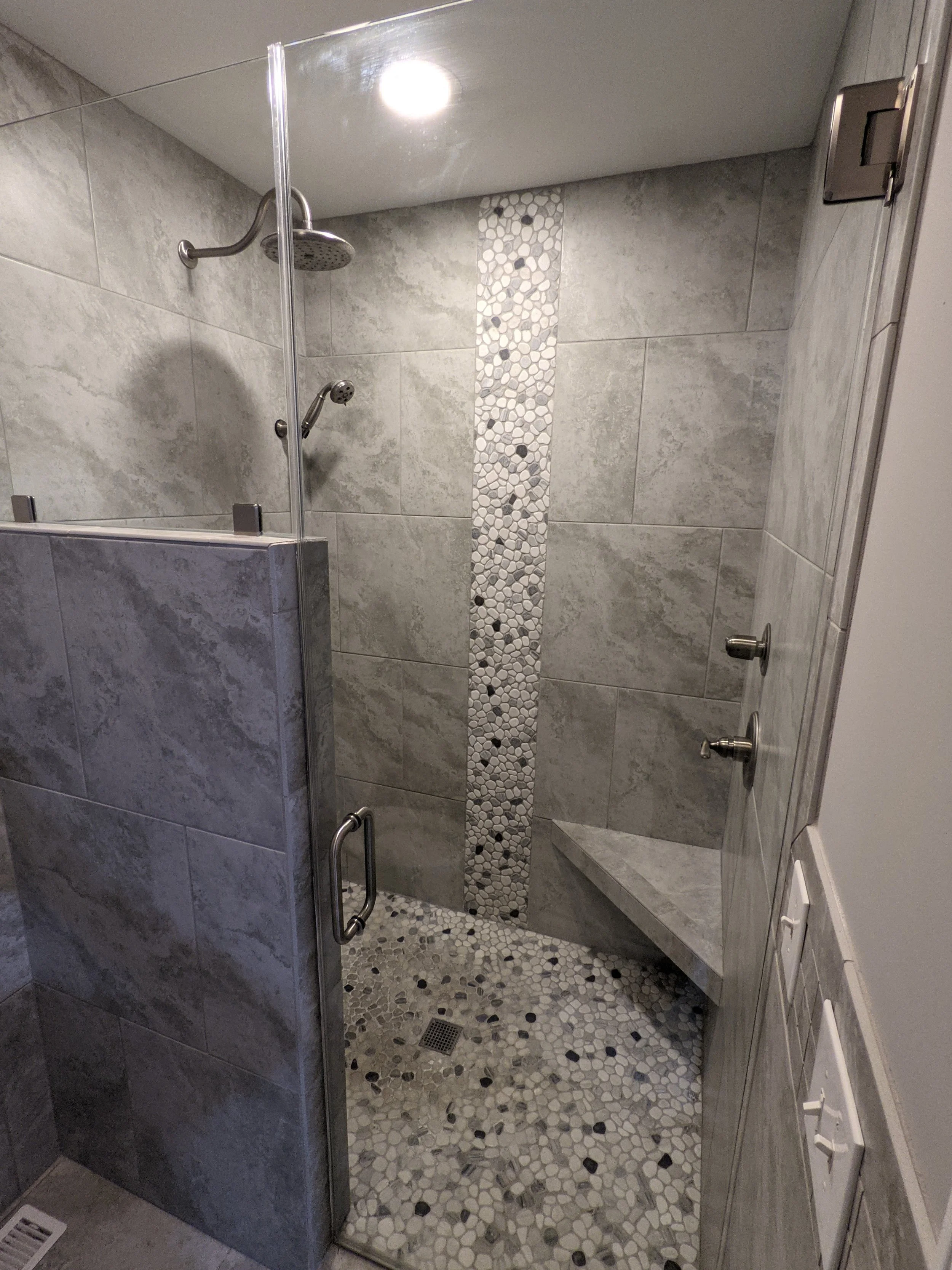 View of a modern walk-in shower with gray tiles, pebble mosaic floor, a vertical strip of pebble mosaic, a built-in corner bench, and two showerheads, enclosed with a glass door.