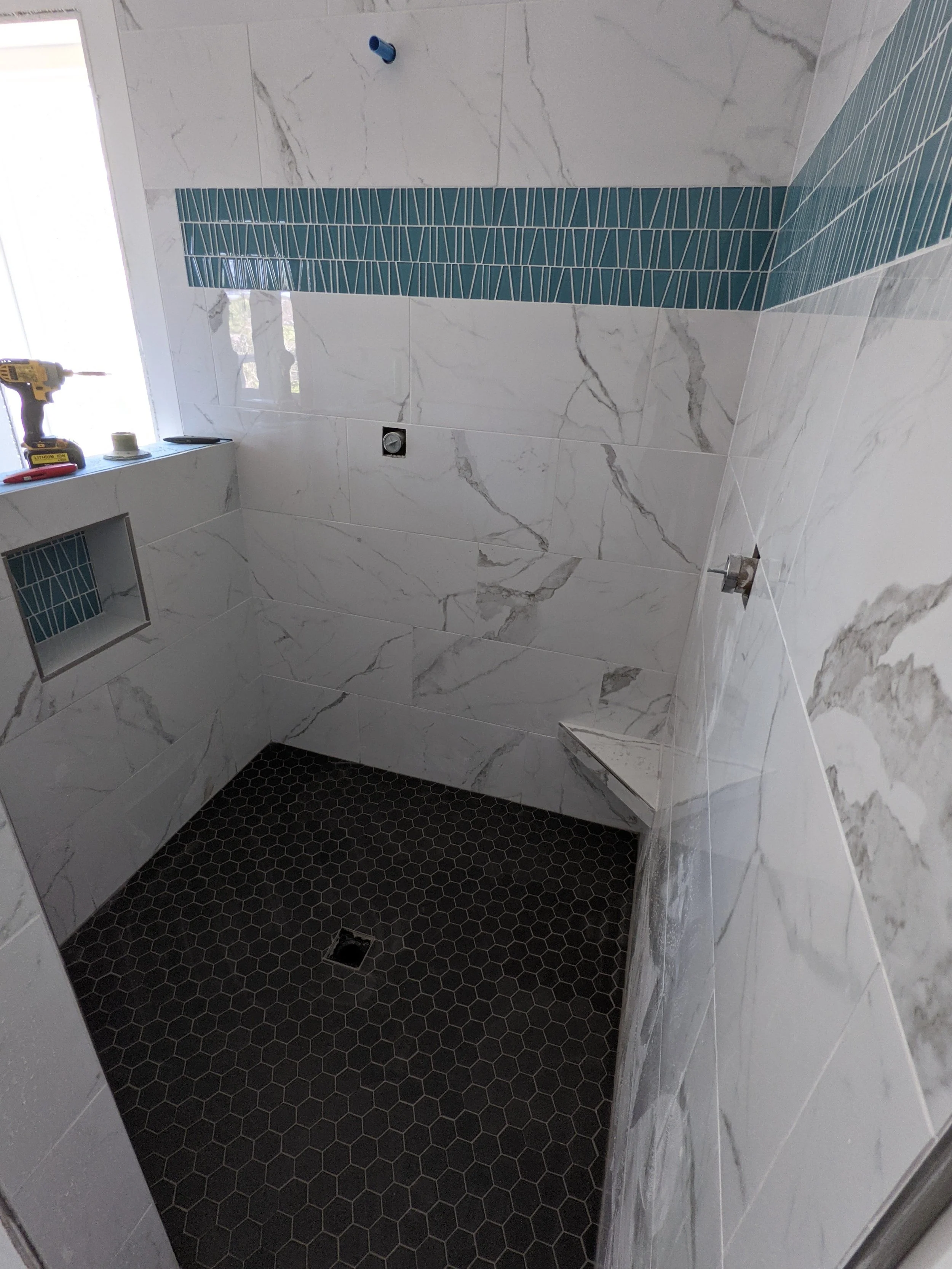 Empty bathroom shower with white marble tiles on the walls, a blue glass tile accent strip, dark hexagonal tile on the floor, and a window with construction tools on the sill.