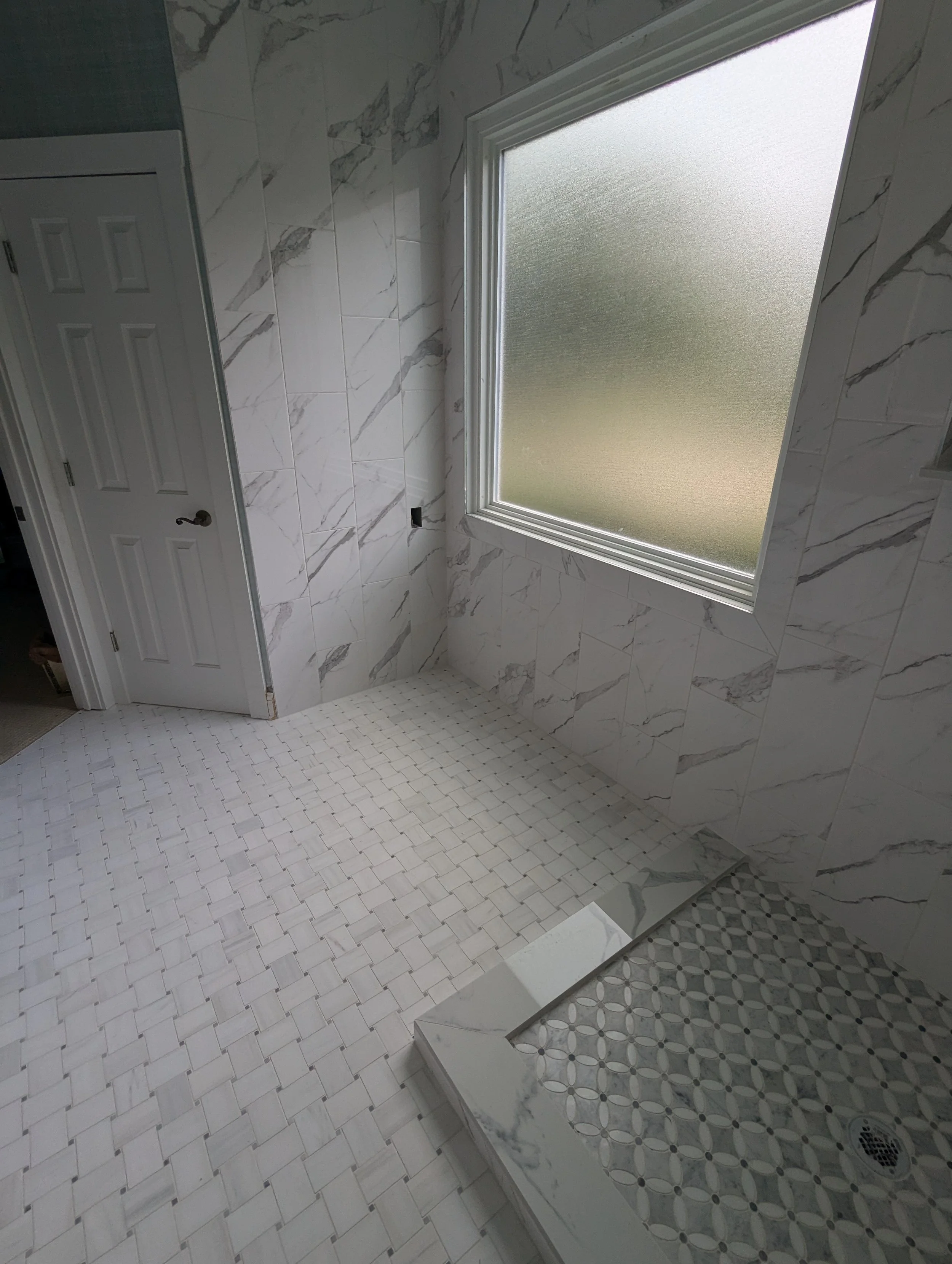 A bathroom with white marble tile walls, a frosted window, a door, and a shower area with patterned tile flooring.