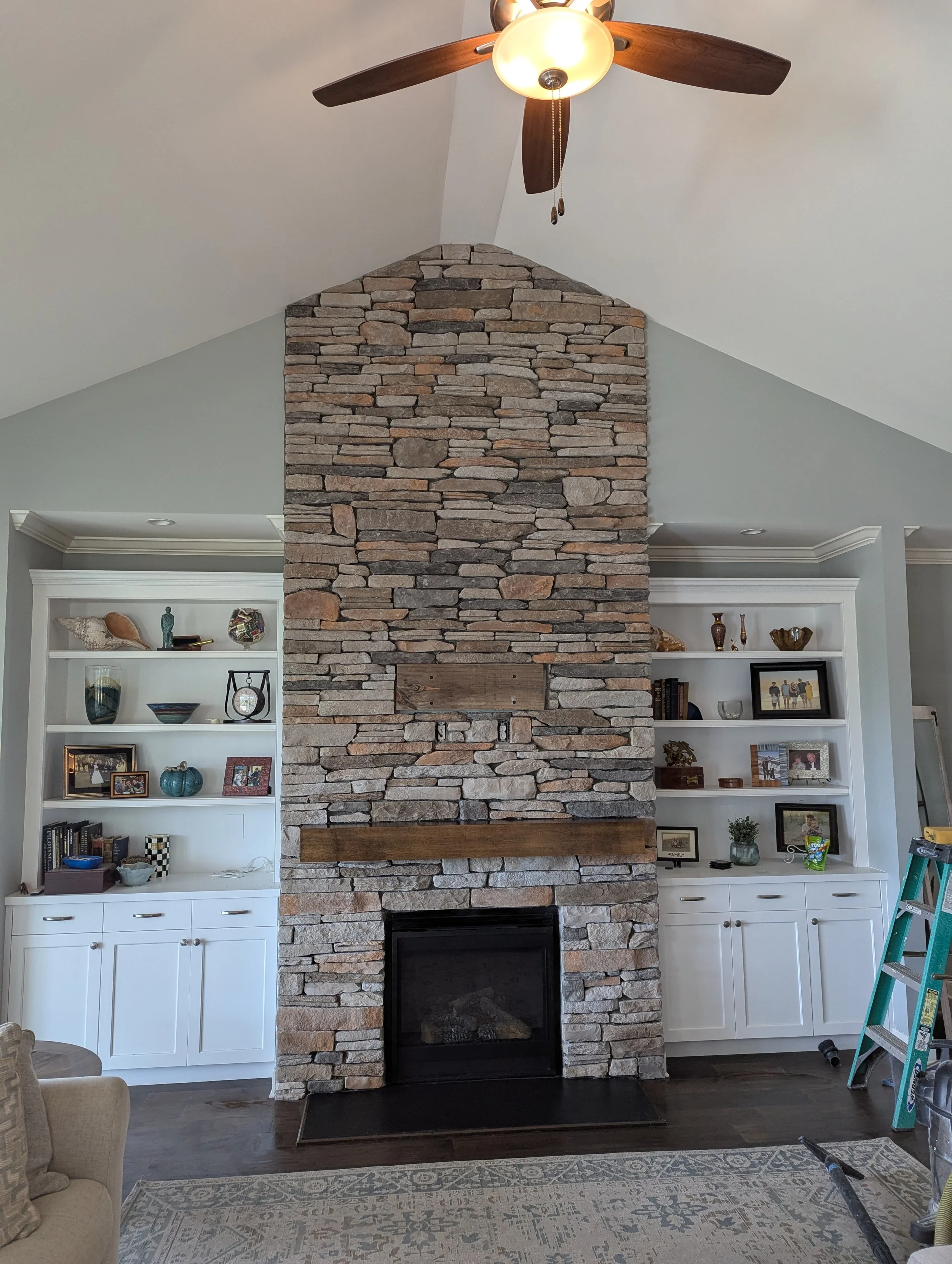 Living room with a tall stone fireplace, white built-in shelves on each side, a ceiling fan, and a ladder on the right side.