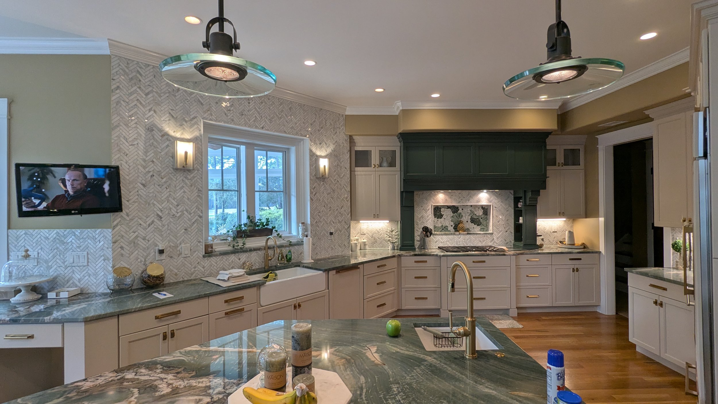 Modern kitchen with white cabinetry, green accents, marble backsplash, hardwood floor, and gold hardware. Features an island with a sink, a window above the sink, a mounted TV, and contemporary lighting fixtures.