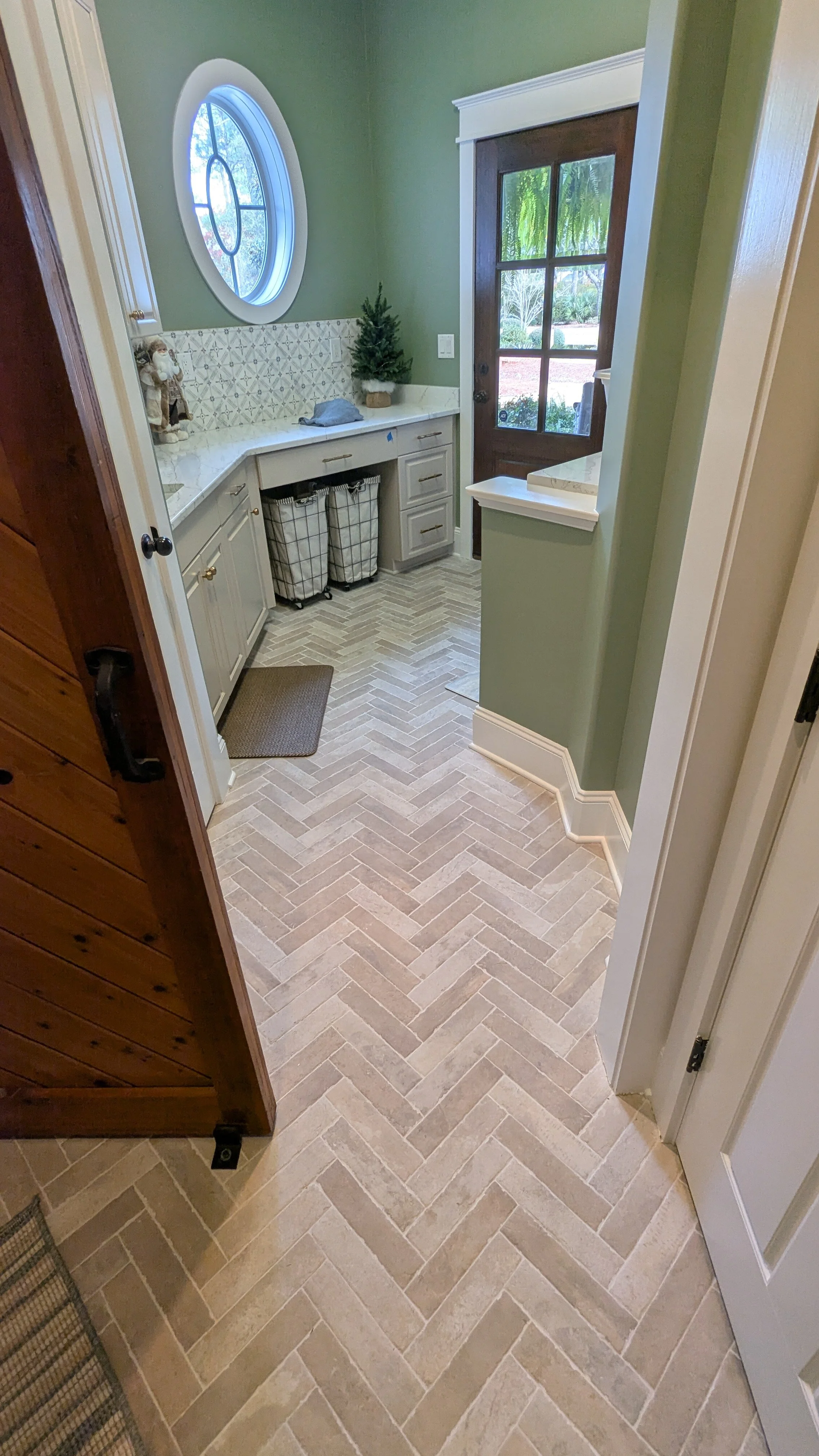 Entryway with a front door, small countertop with a decorative tree, and a window with a decorative grille. The floor features beige and light brown herringbone tile, and there are small laundry baskets under the countertop.