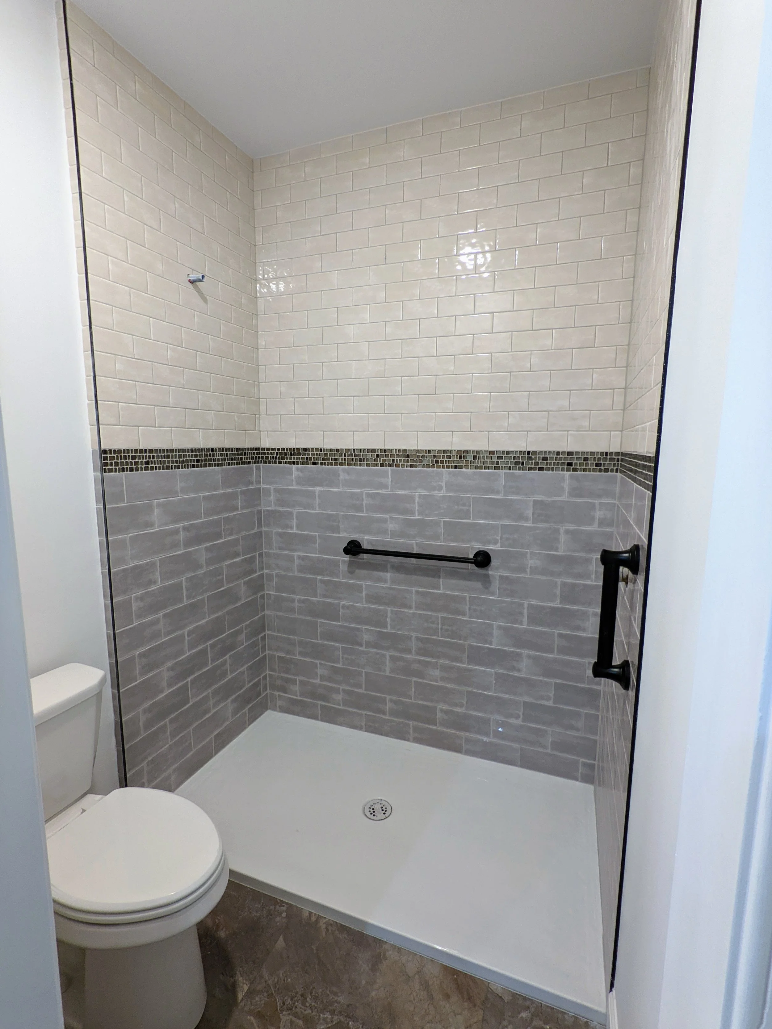 A walk-in shower with beige and gray tiled walls, black grab bars, a drain in the floor, and a toilet beside it.