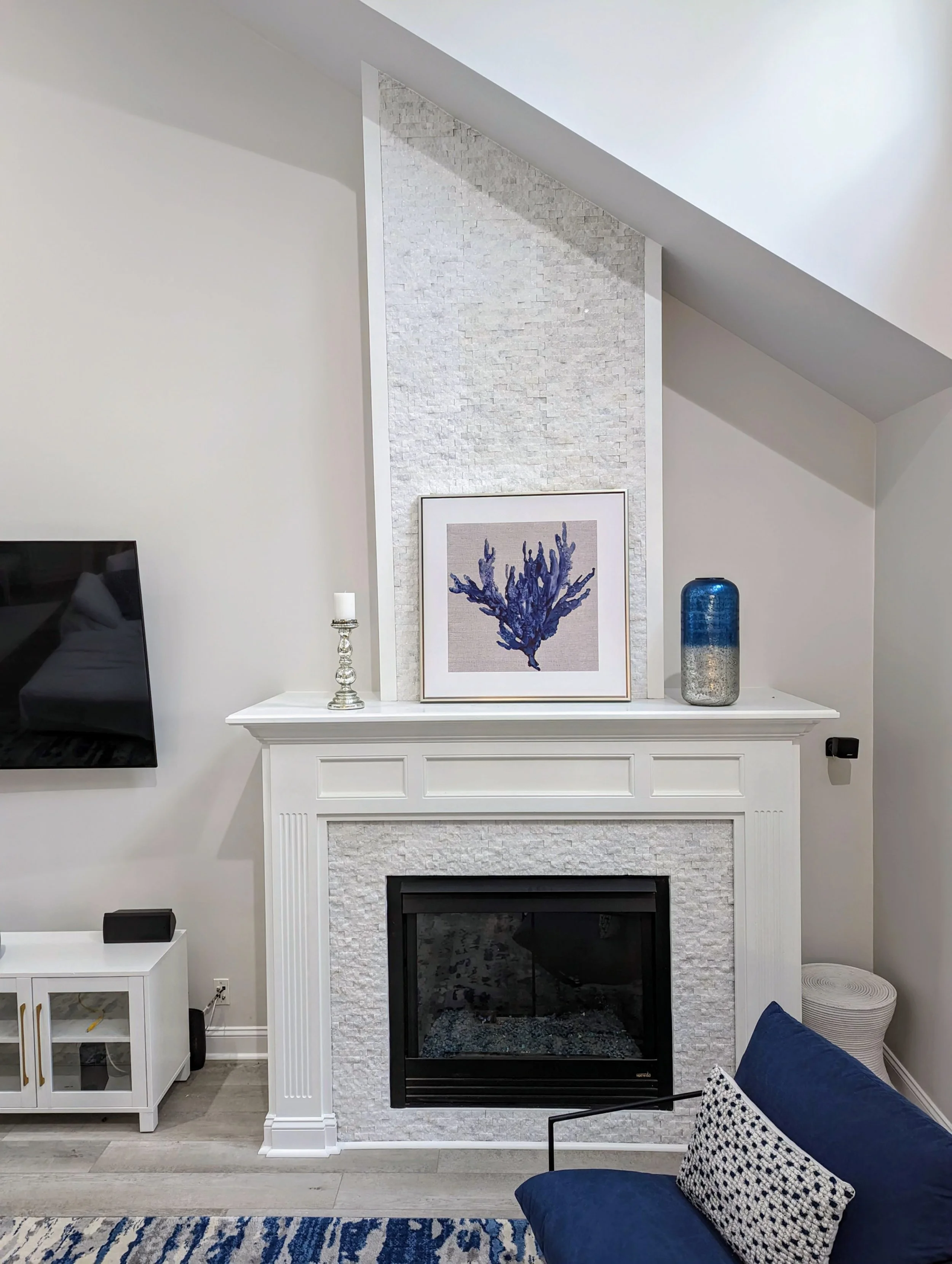 Living room with white fireplace, framed blue coral artwork on the mantel, large blue and silver vase, and a crystal candlestick. Part of a navy sofa with a black and white pillow is visible in the foreground.