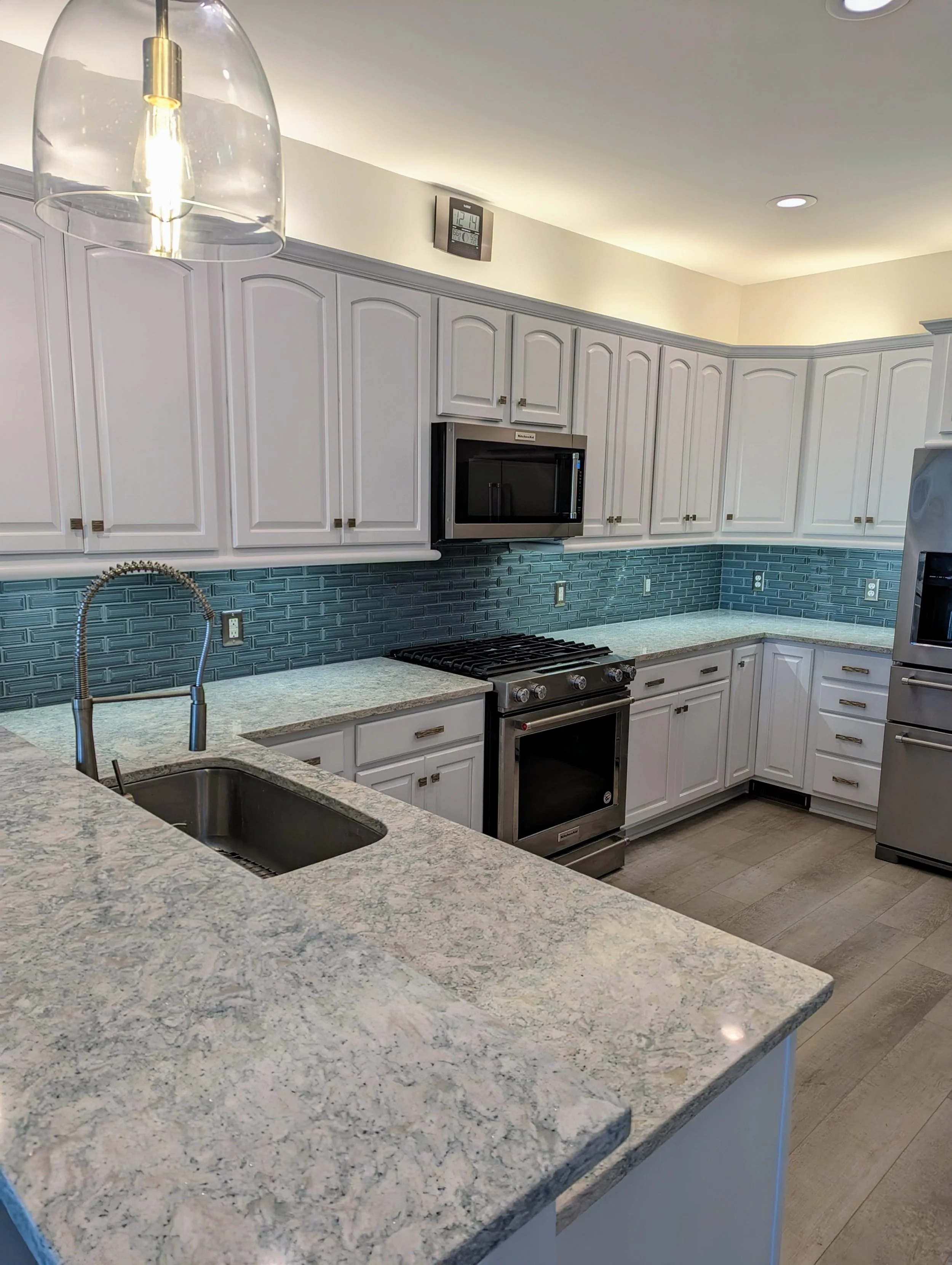 Modern kitchen with white cabinets, blue subway tile backsplash, granite countertops, stainless steel appliances, including a microwave, stove, and refrigerator. There is a kitchen island with a sink and a pendant light overhead.