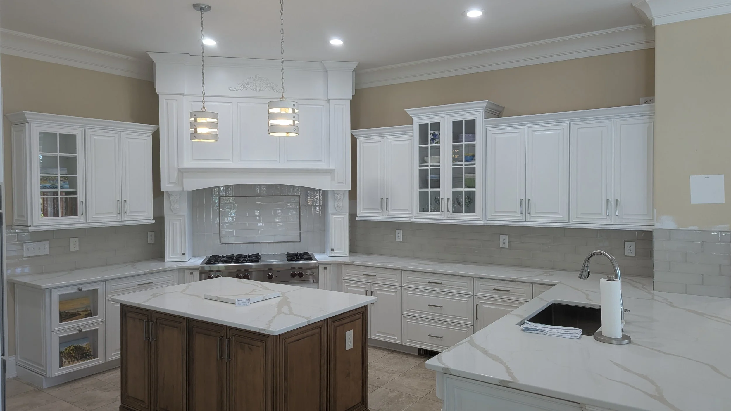 A bright kitchen with white cabinets, a white marble island, and a wooden corner cabinet. There are stainless steel appliances and a tiled backsplash behind the stove. Recessed ceiling lights and two pendant lights hang above the island.