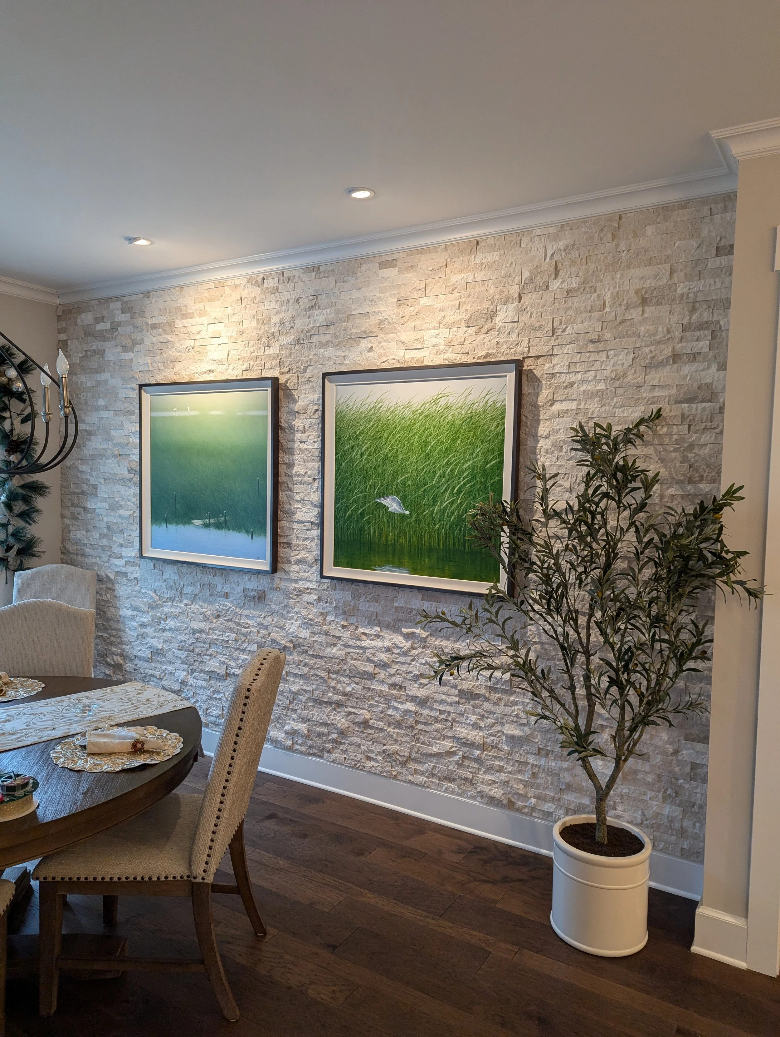 Interior of a dining room with a white textured stone wall, two framed nature photographs of green grass and water, a potted olive tree, and a dark wooden dining table with beige upholstered chairs.