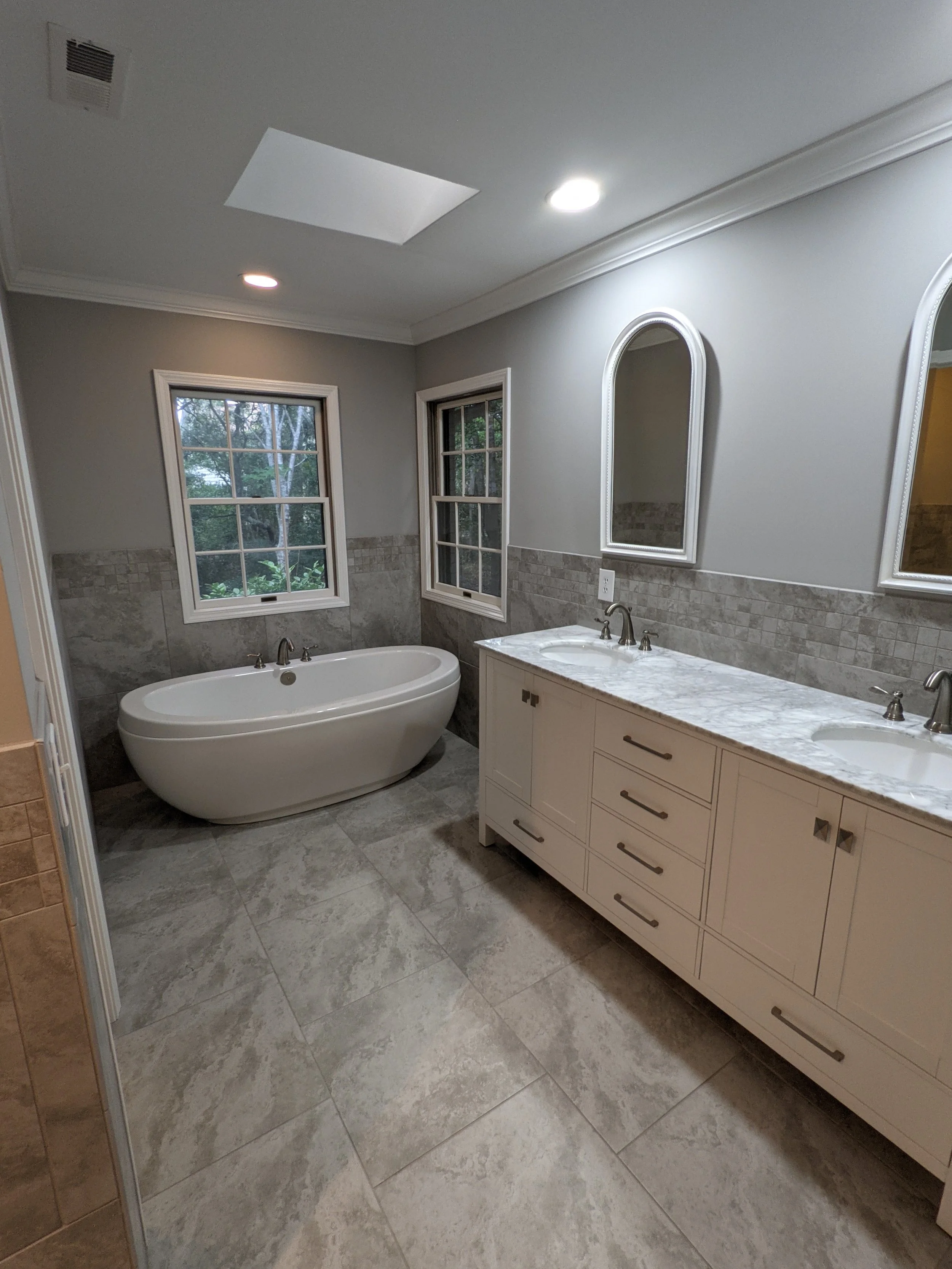 A modern bathroom featuring a freestanding bathtub, double sink vanity with marble countertop, large mirrors, tiled floors, and two windows with a view of ? outside.