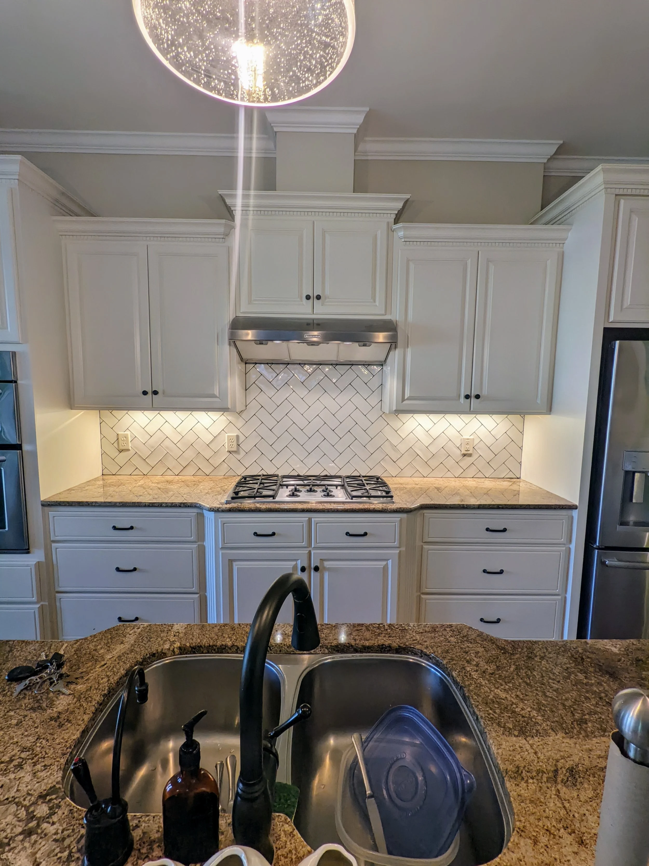 A kitchen with white cabinets, a granite countertop, a tiled backsplash, and a stainless steel range hood above a gas stove. The image is taken from the perspective of someone standing at a kitchen island with a sink.